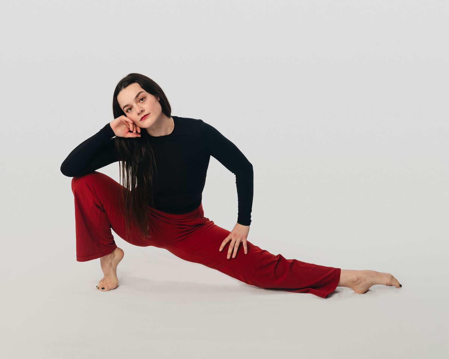 Dancer in a deep lunge wearing red pants and a black top, looking confidently at the camera in a studio setting. Dance portrait photography.