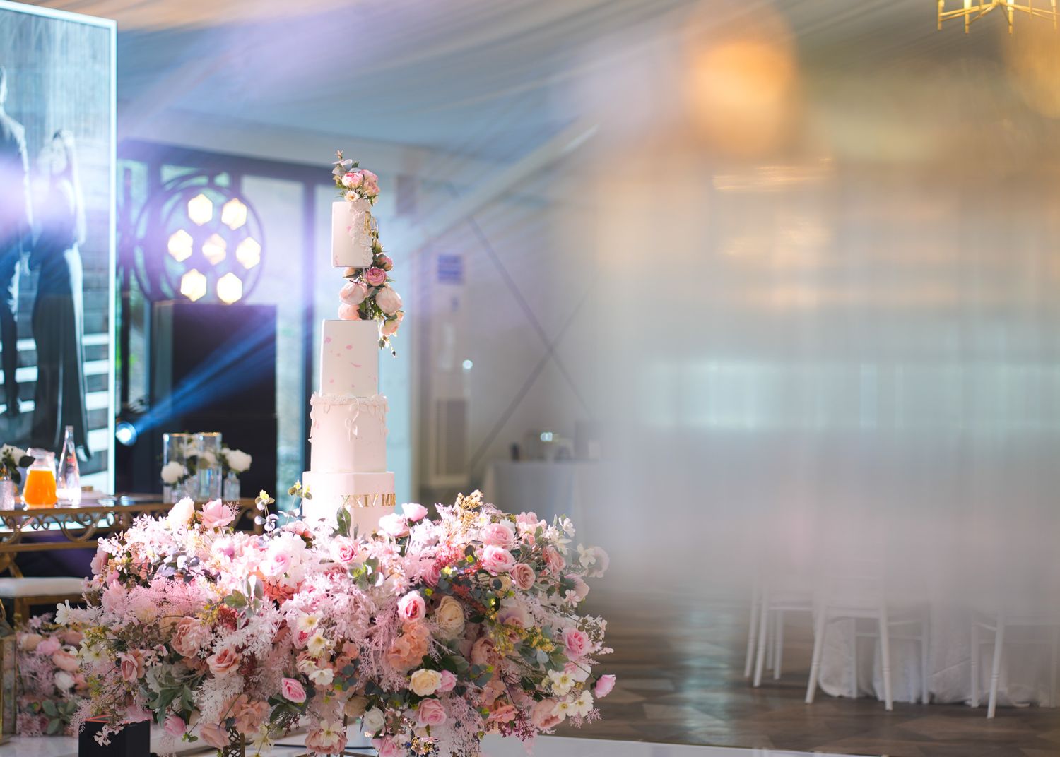 Elegant white and pink tiered wedding cake adorned with fresh flowers, displayed on a table surrounded by lush pink floral arrangements.