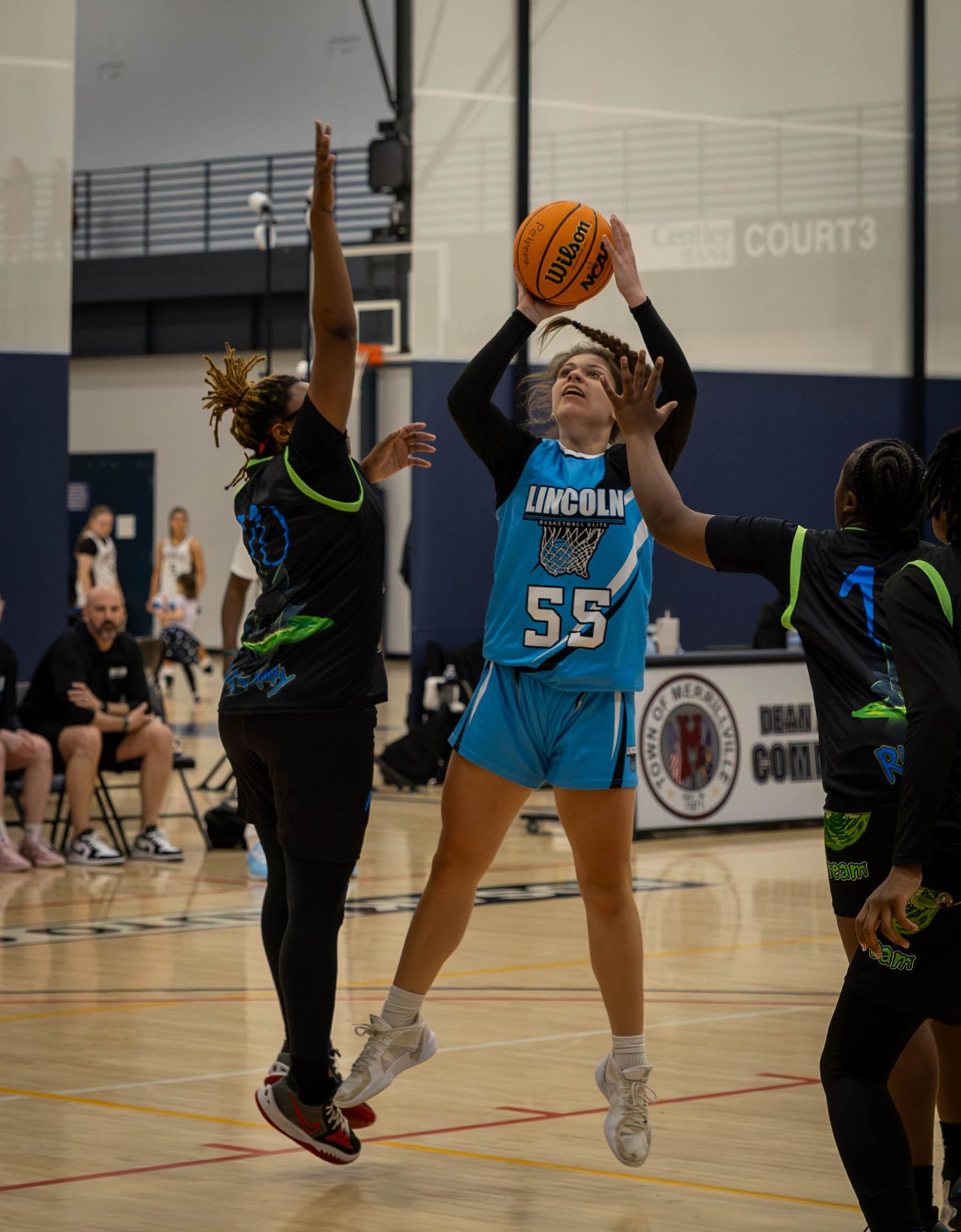 Basketball player in light blue uniform jumps for a shot while two defenders in dark uniforms attempt to block.