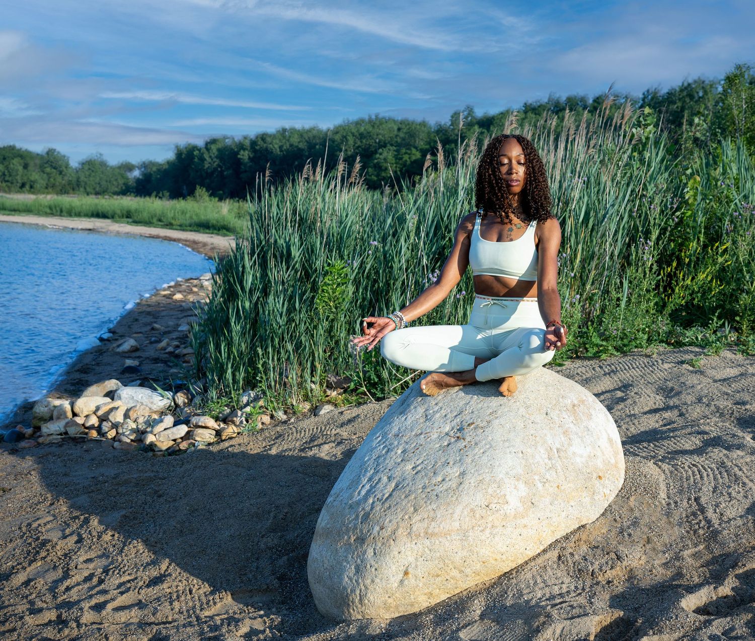 Someone practices meditation in lotus pose on a large boulder by a serene lake with tall reeds and blue sky.