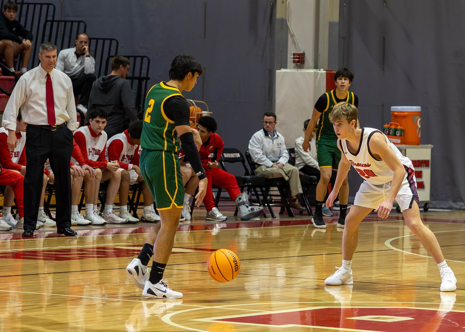 High school basketball players compete on indoor court, green uniform player dribbles while defender guards in red and white.