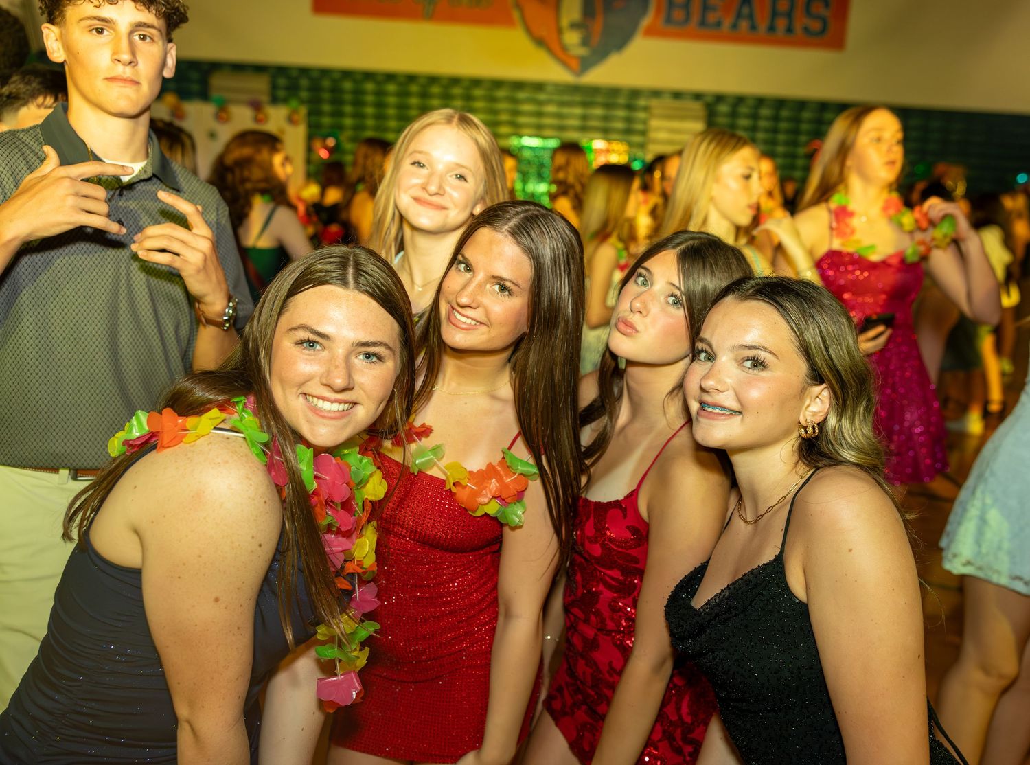 Group of teenage girls in red dresses and floral leis smiling at a school dance in a gym.