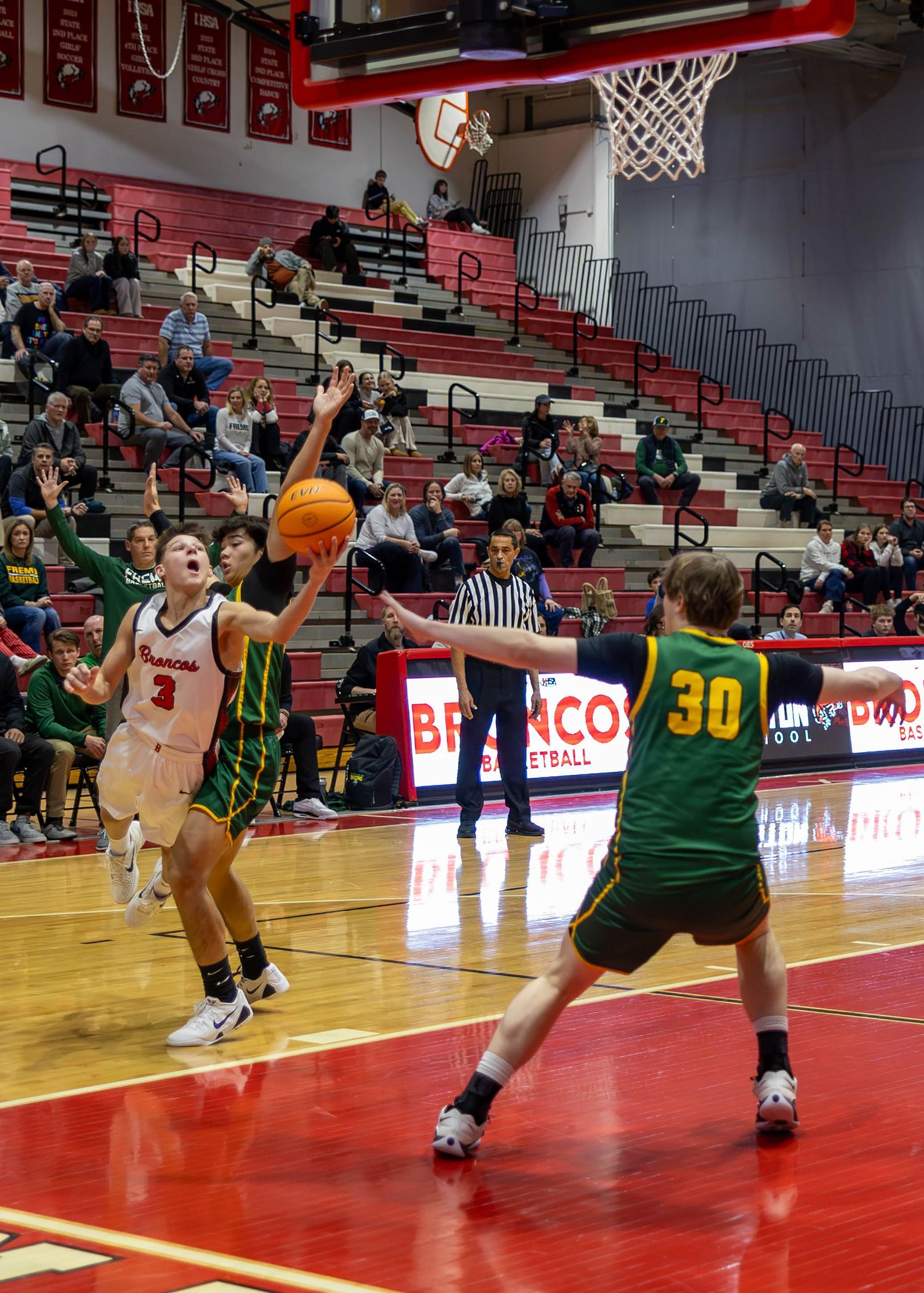 High school basketball player in white uniform attempts a layup against defender in green number 30 jersey in indoor gym.