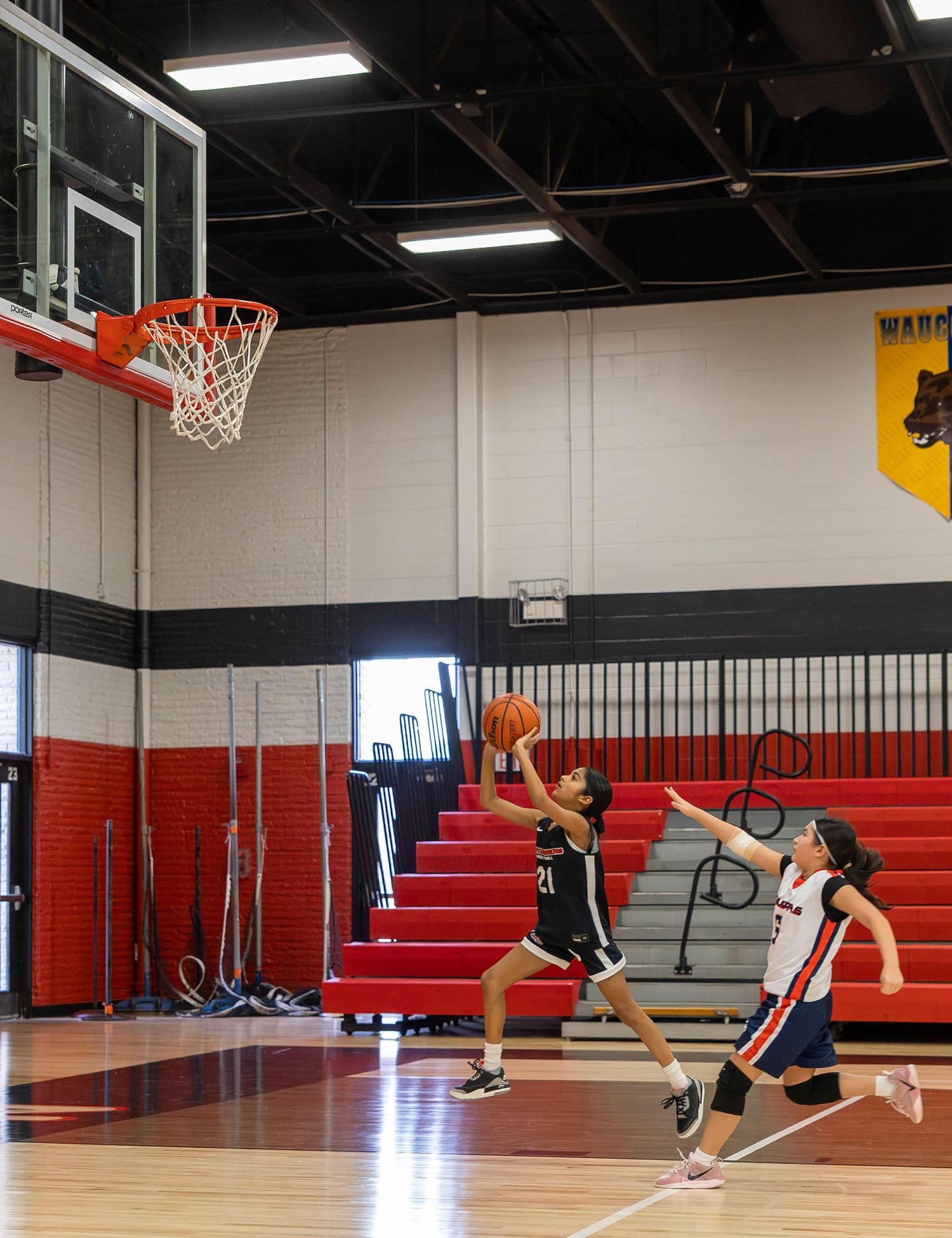Two youth basketball players compete in a gym, one in black attempting a layup while a defender in white reaches to block.
