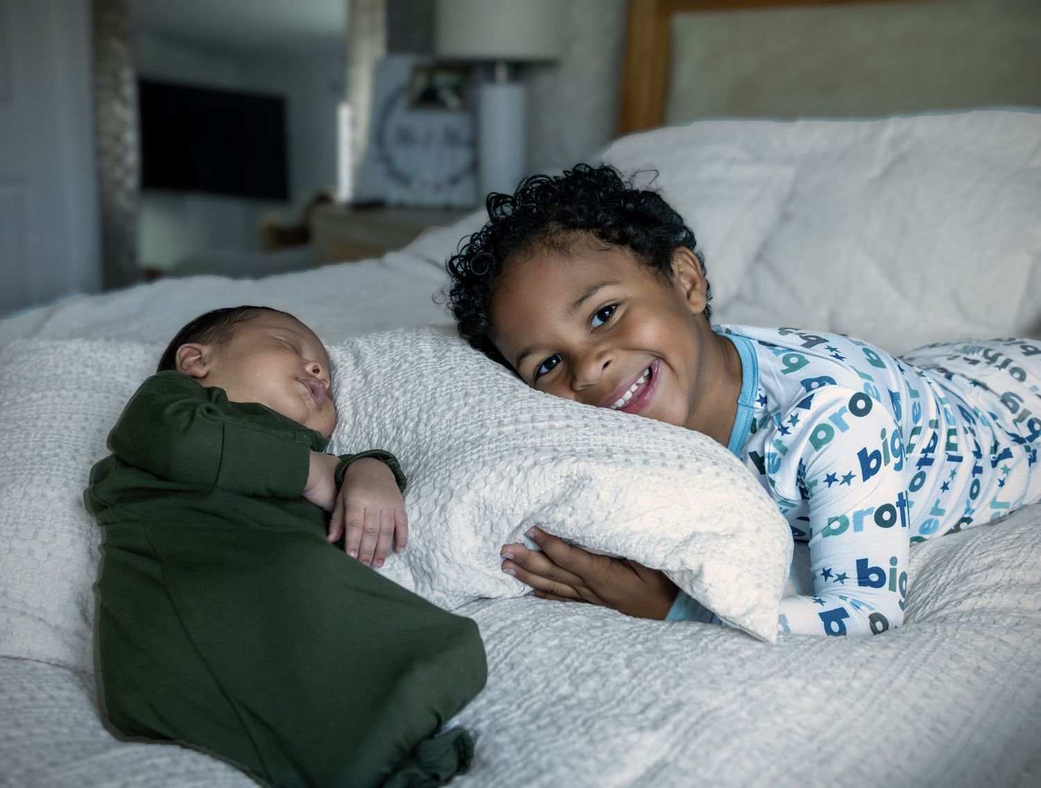 Smiling toddler in pajamas lying on a bed next to a sleeping newborn swaddled in a green blanket.