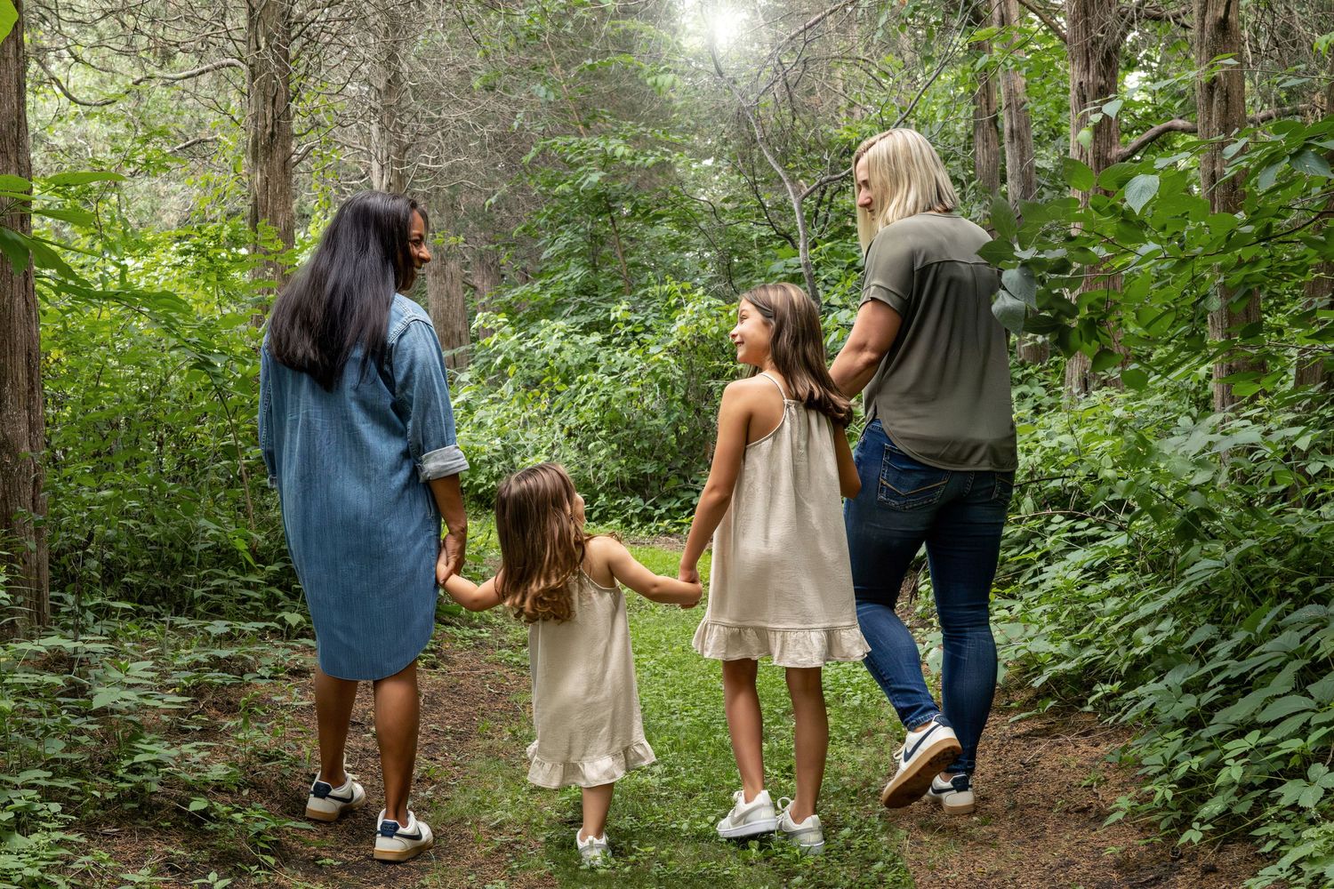 A family walks together holding hands on a forest trail surrounded by lush green foliage.