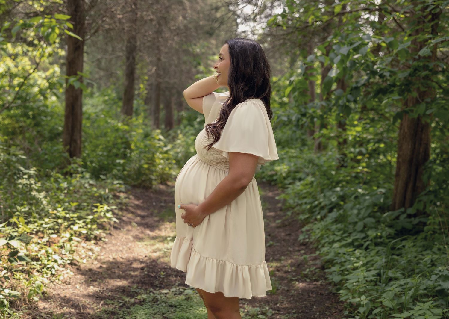 Pregnant woman in cream dress holding belly on forest path.