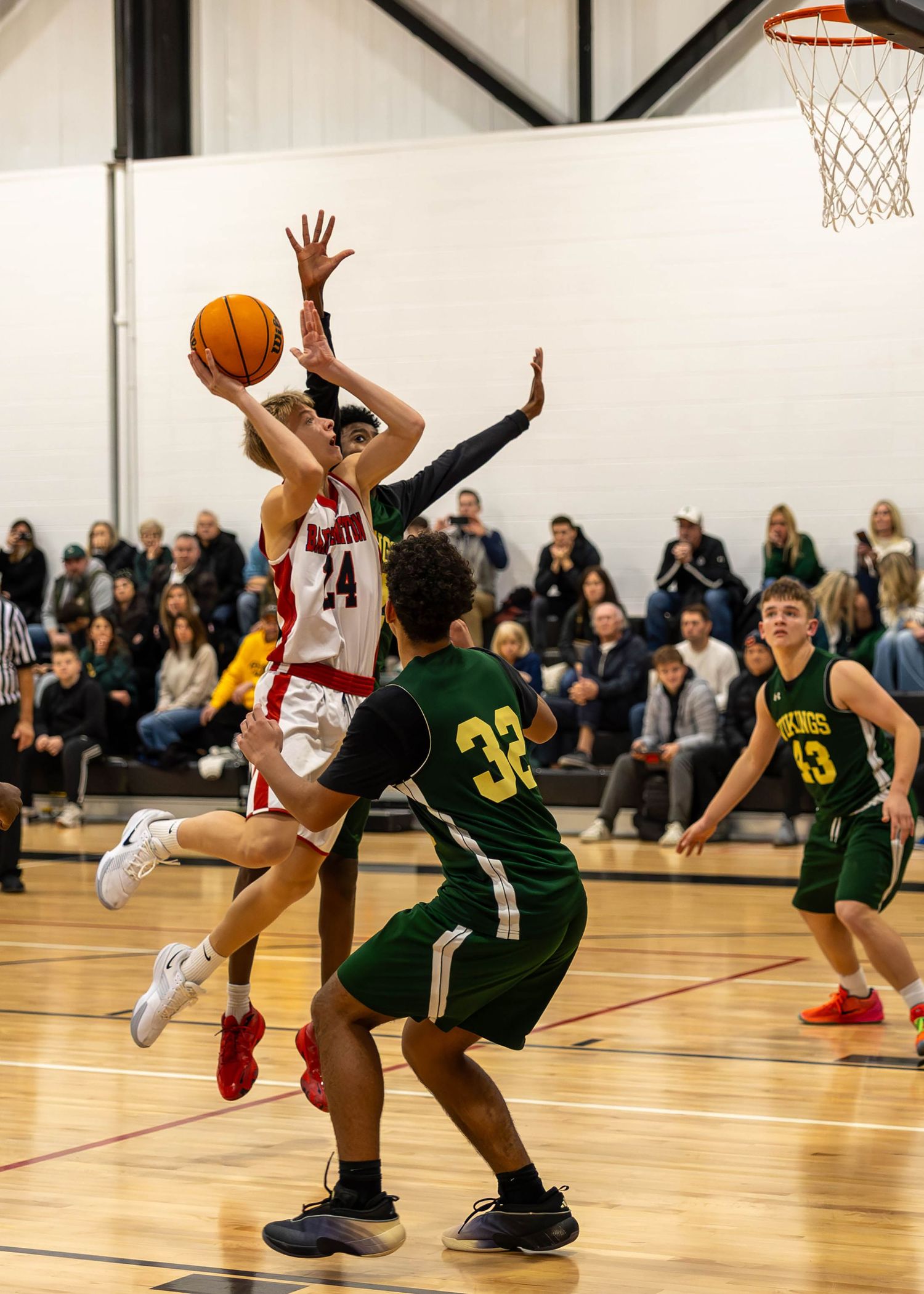 High school basketball player in red uniform drives to the basket against green-uniformed defenders in a packed gymnasium.