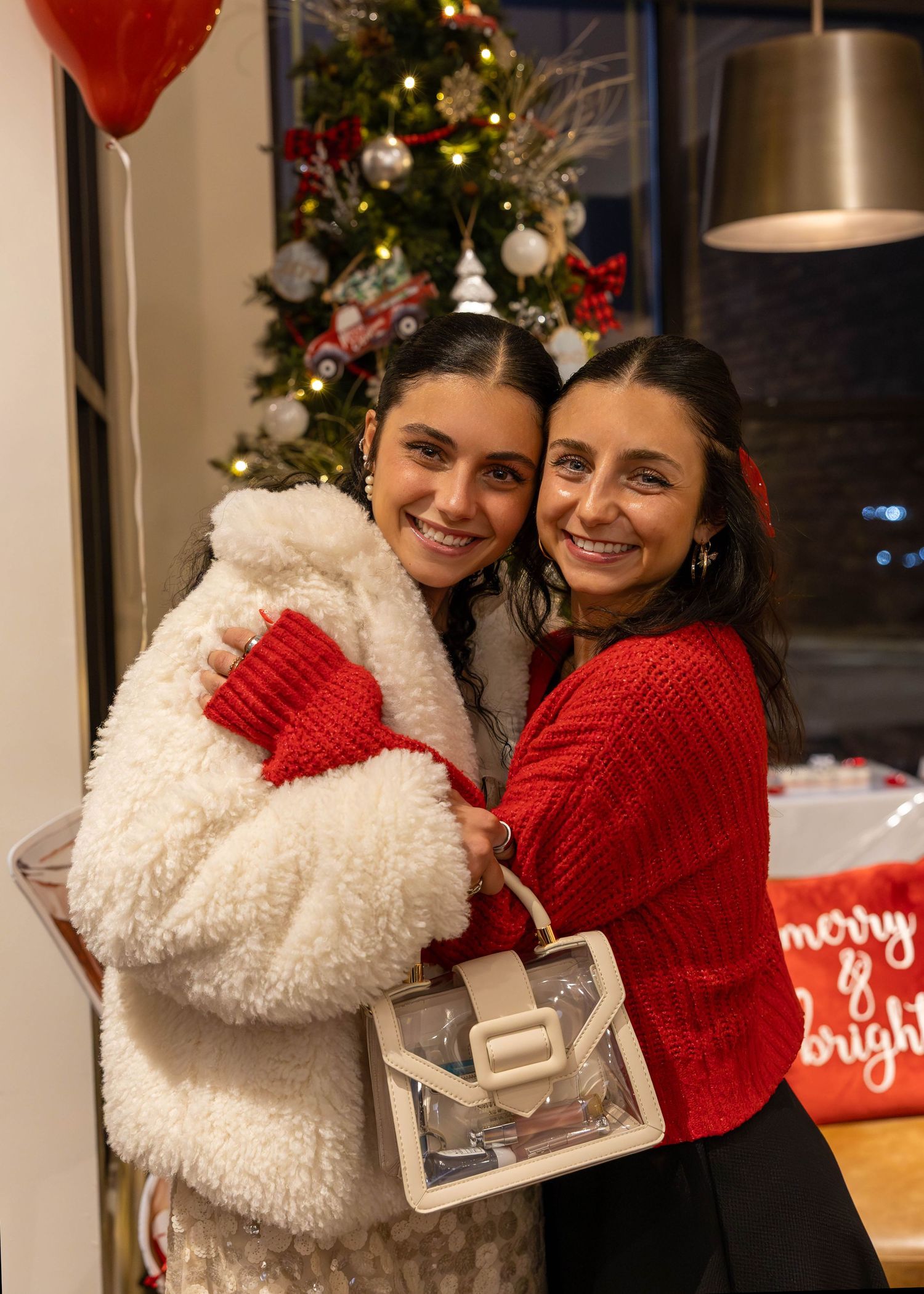 Two women hugging and smiling in front of a decorated Christmas tree, one wearing a white fluffy coat with red gloves.
