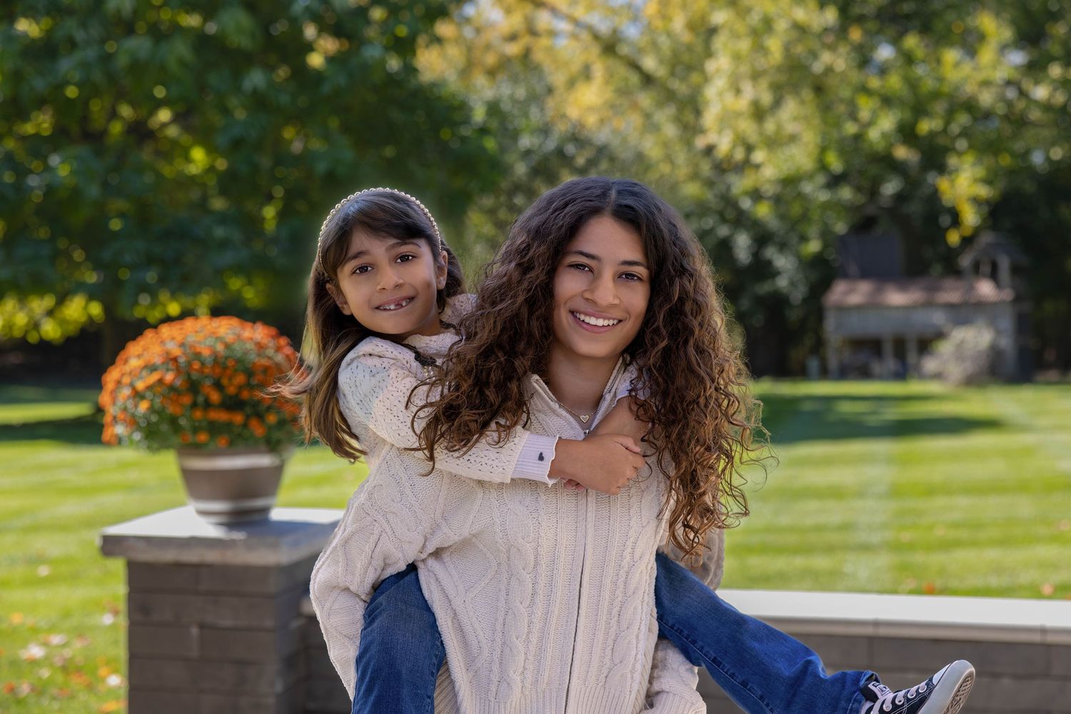 Two smiling girls pose outdoors, older teen giving younger child a piggyback ride in a lush green garden setting.