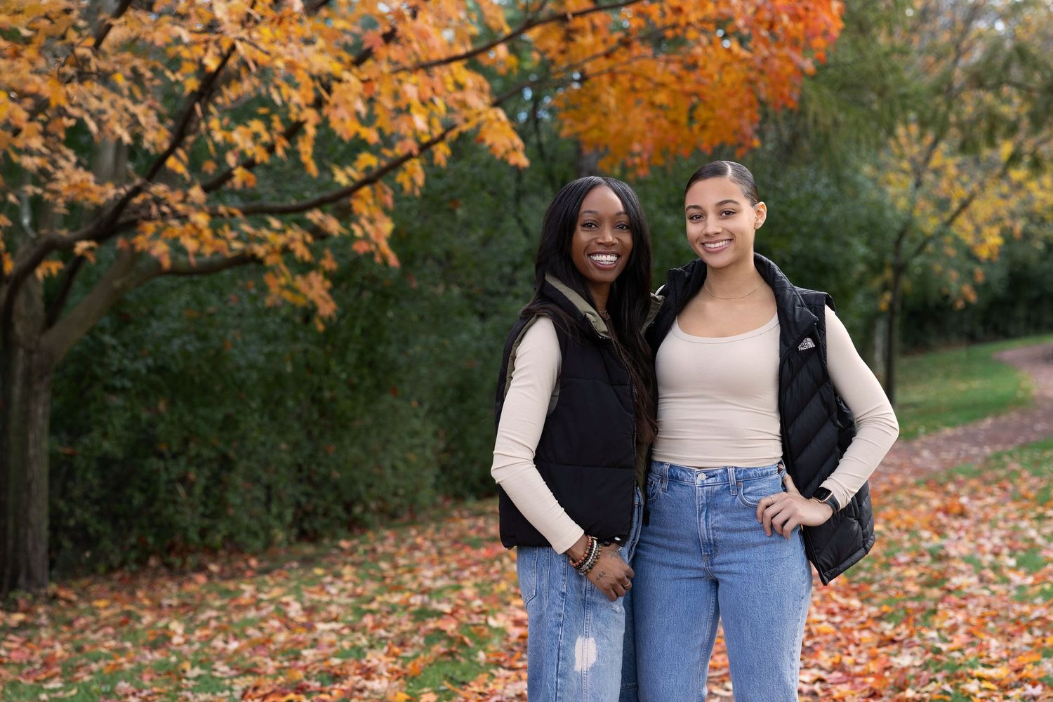 Two young women smiling outdoors in autumn park with colorful orange and yellow fall foliage trees behind them.