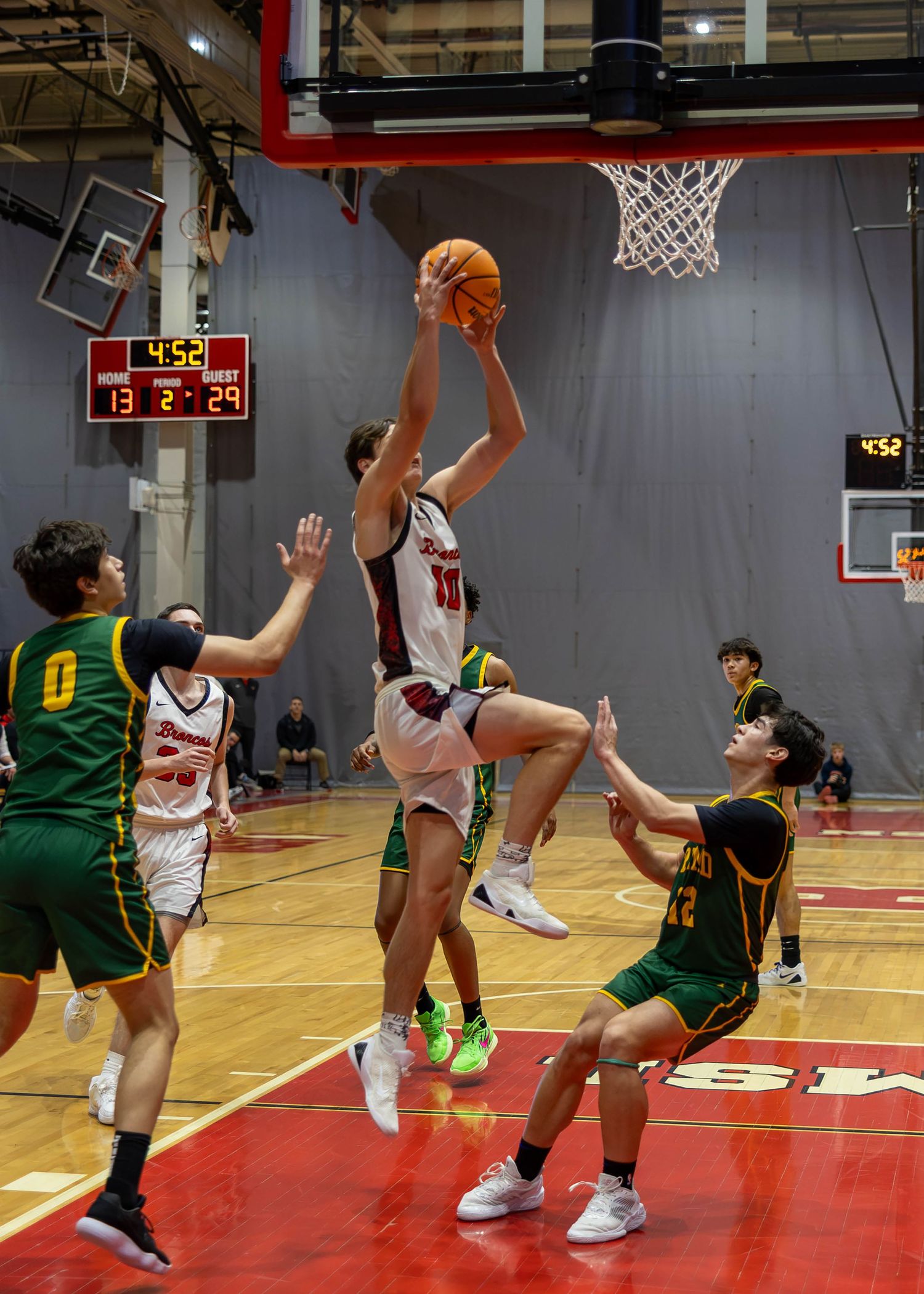 A basketball player leaps for a layup during a high school game, surrounded by opponents in green uniforms on a red court.
