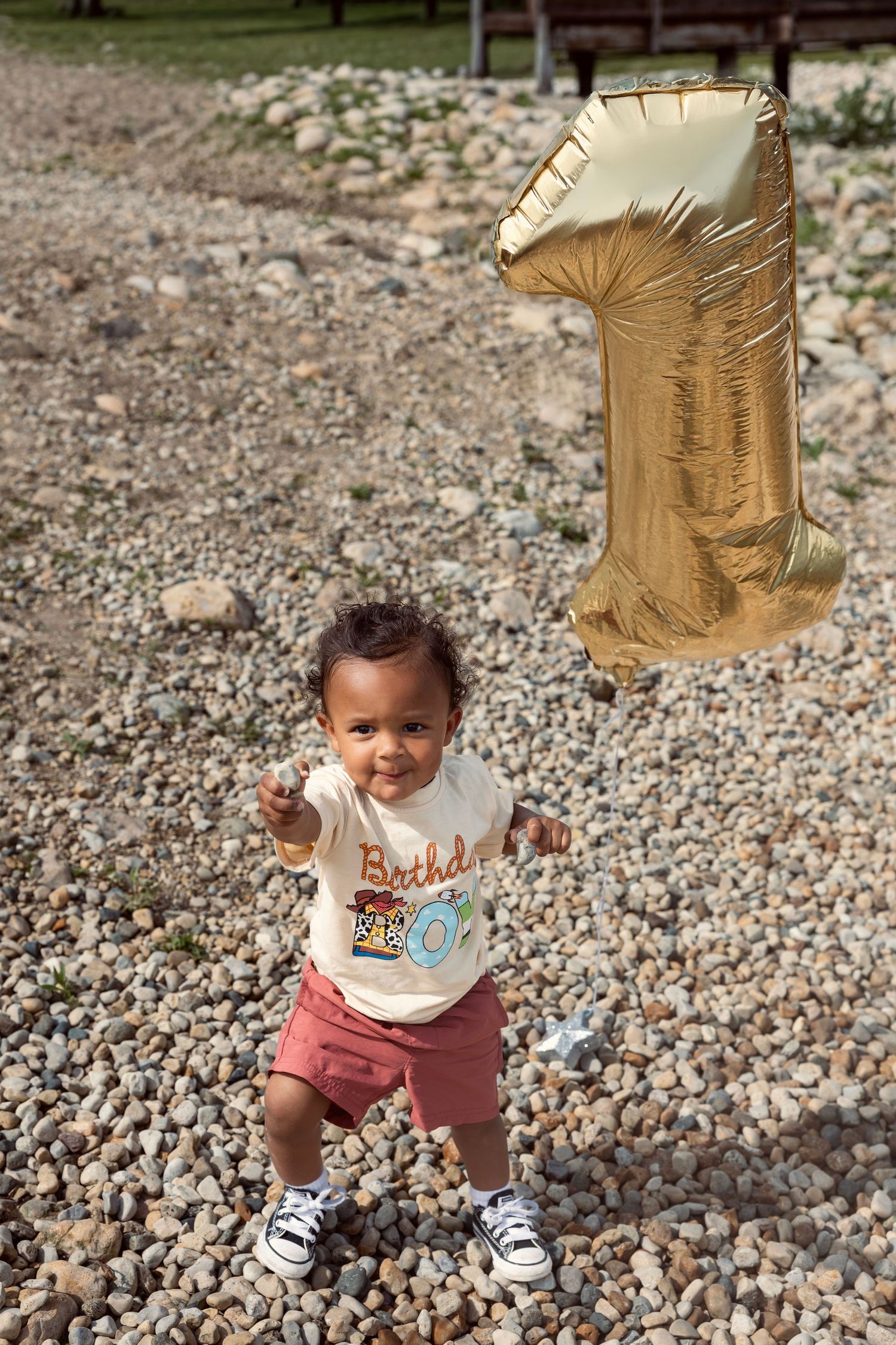 Toddler in a birthday shirt holds a gold number 1 balloon on a rocky outdoor surface, celebrating their first birthday.
