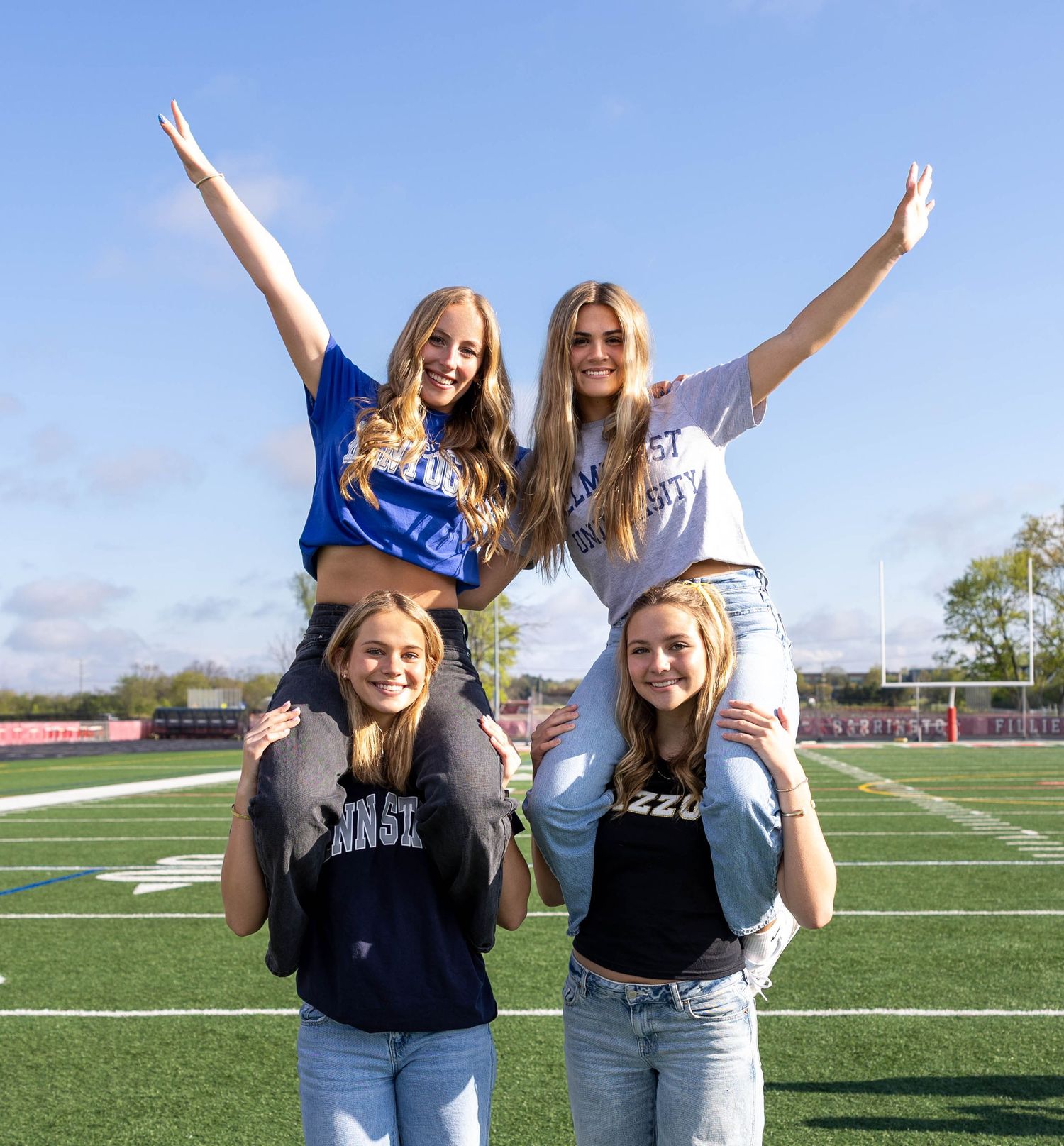 Four cheerleaders pose on a football field, two on shoulders with arms raised, wearing college spirit shirts.