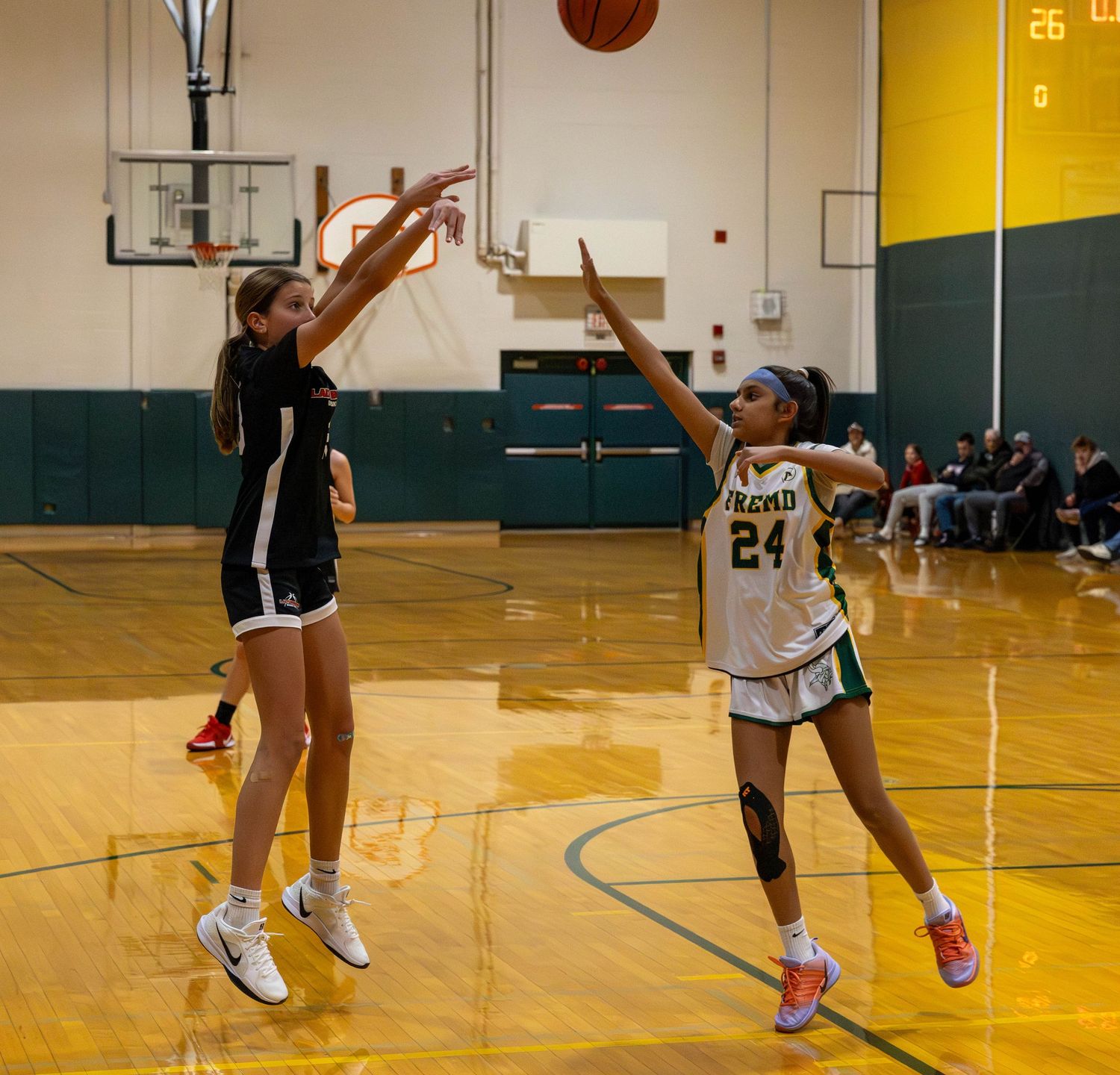 Two female basketball players compete during an indoor game, one in black shooting over a defender in green jersey number 24.