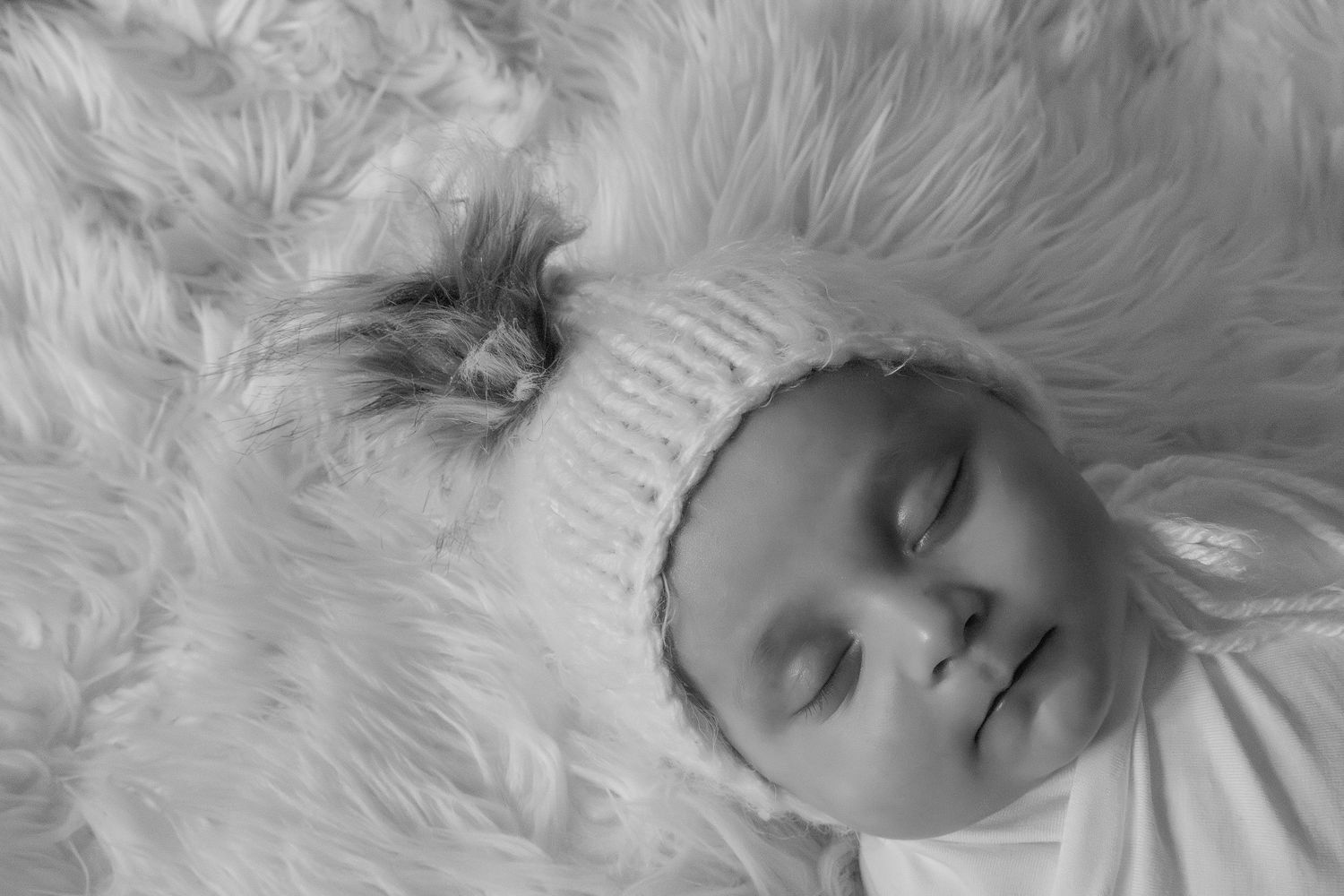 Black and white photo of a sleeping newborn baby wearing a knit bonnet on a fluffy white fur rug.