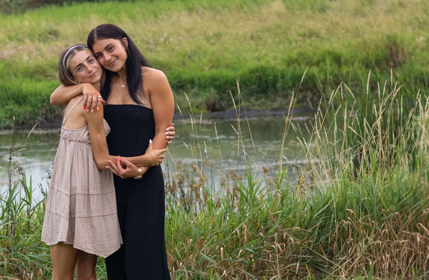 sisters embrace while standing near water with tall grass in summer setting.