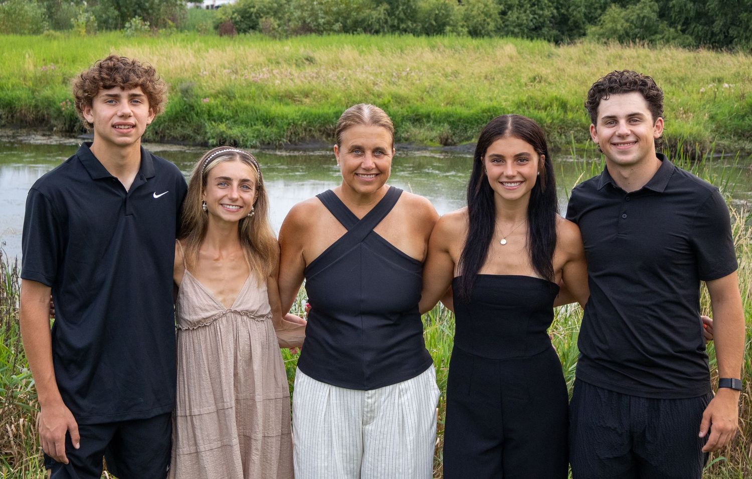 A family of five people pose together outdoors near a pond on a sunny summer day.