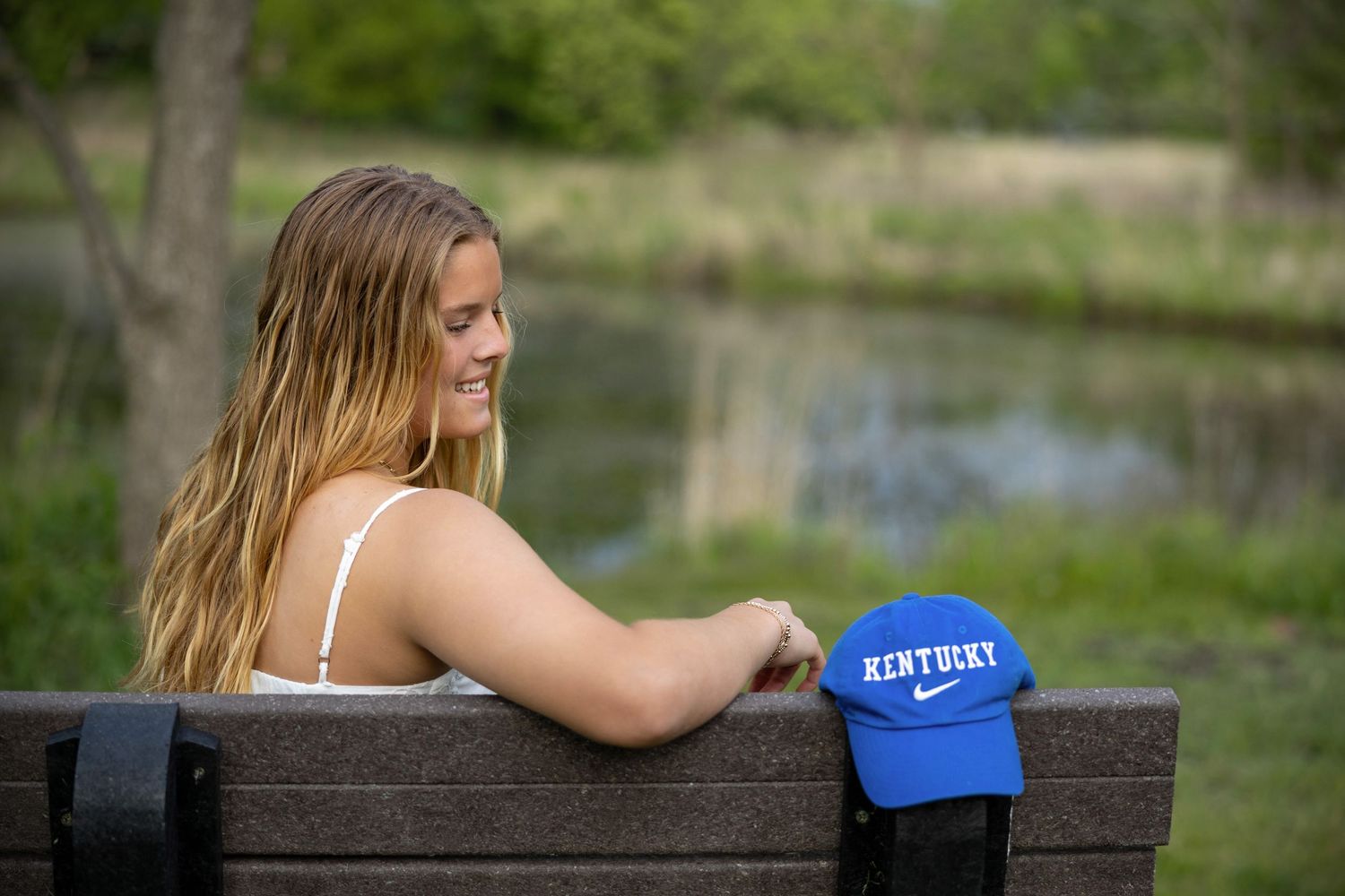 Someone in a white top sits on a wooden bench near a lake with a Kentucky blue baseball cap beside them.