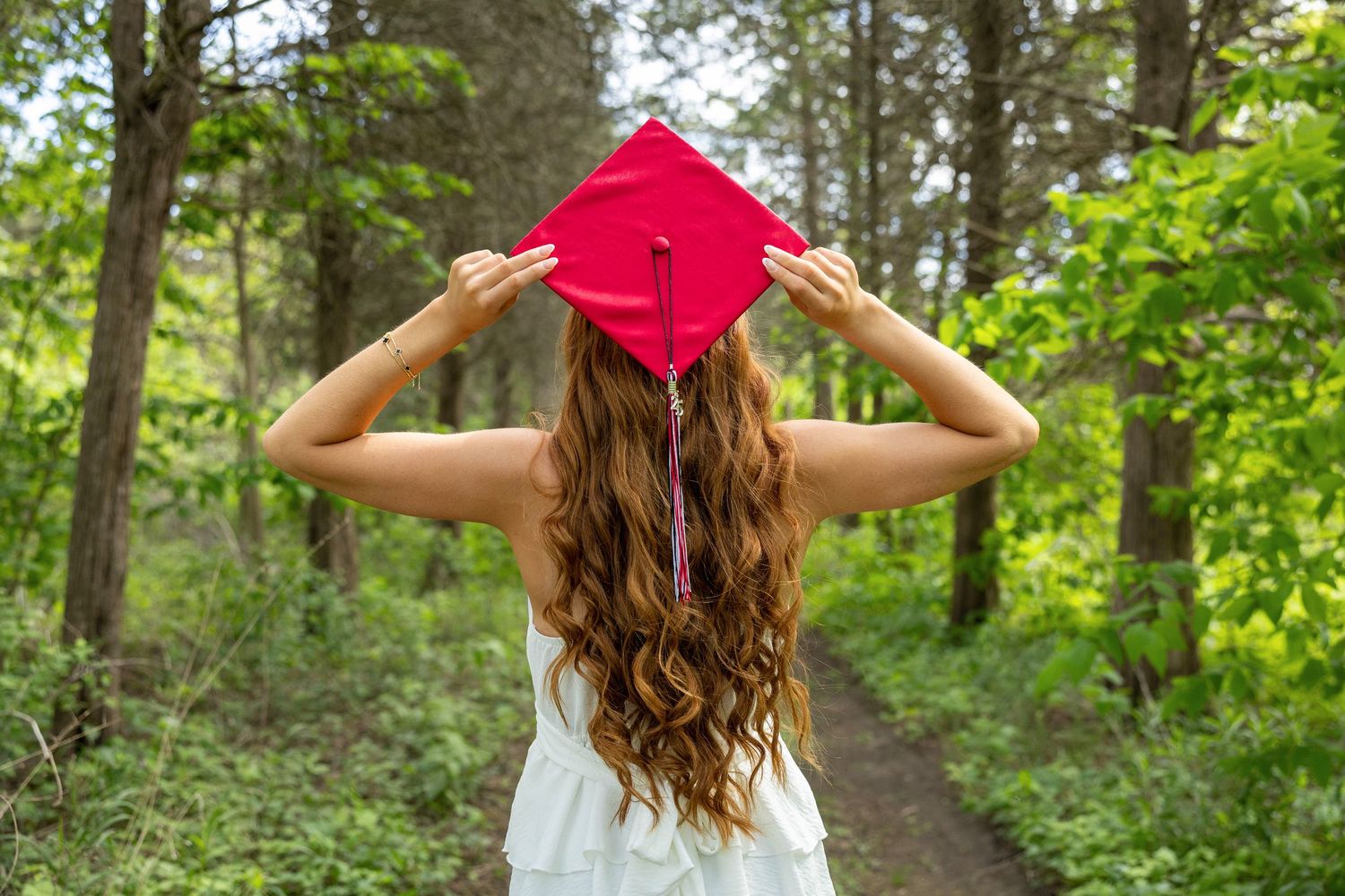 Graduate in white dress stands on forest path holding bright red mortarboard cap on a sunny day.