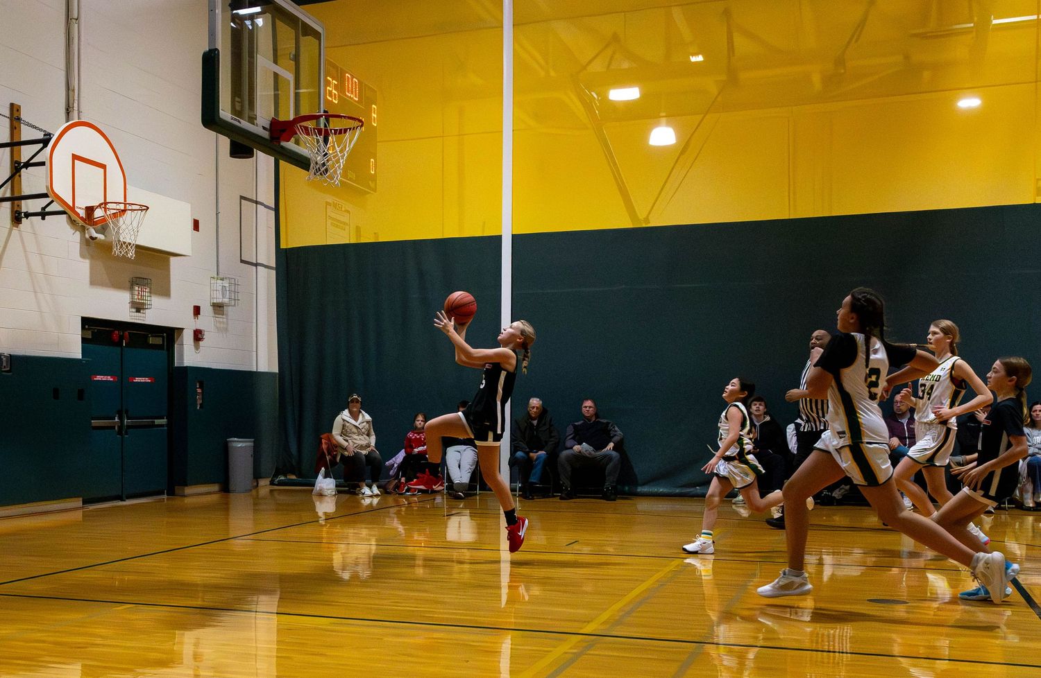 Female basketball player shoots during an indoor game while teammates and opponents run on a green and yellow court.