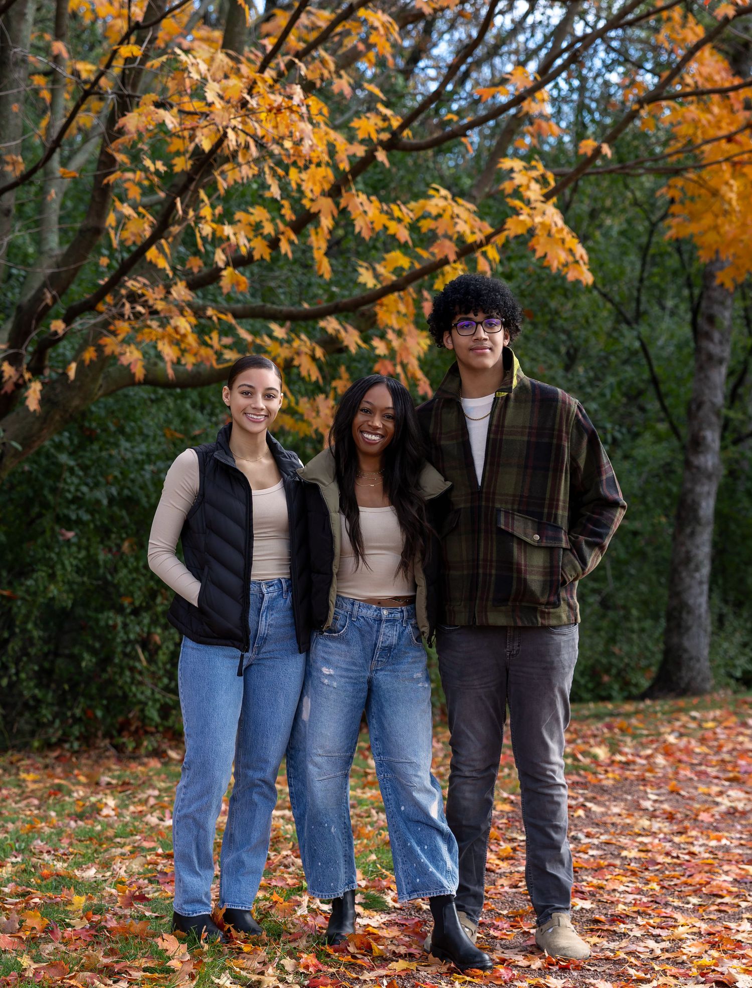 Three friends smiling outdoors in autumn park with colorful orange and yellow foliage, wearing casual fall outfits.