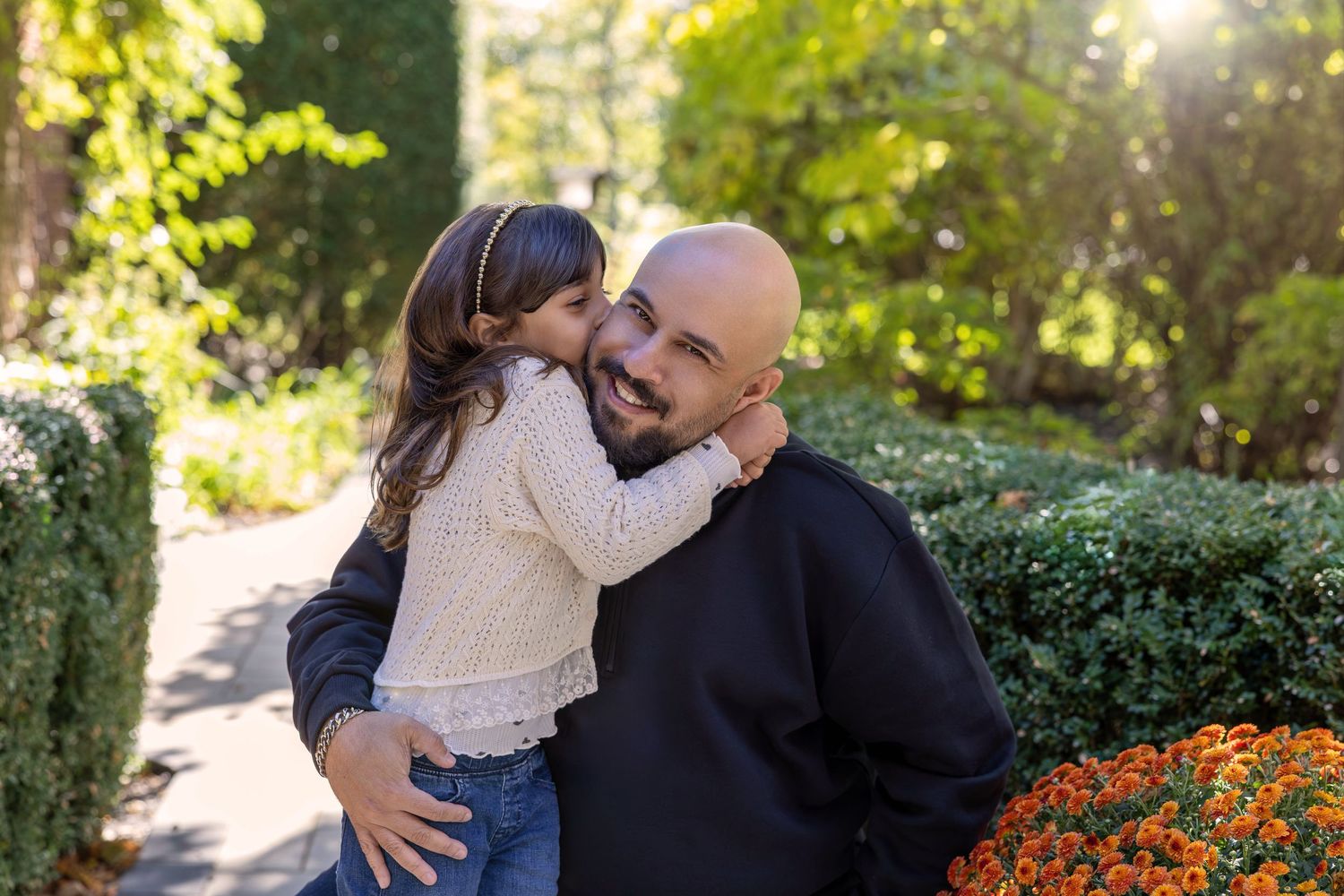 A bald man holds a young girl in a sunny park as she kisses his cheek near orange flowers.