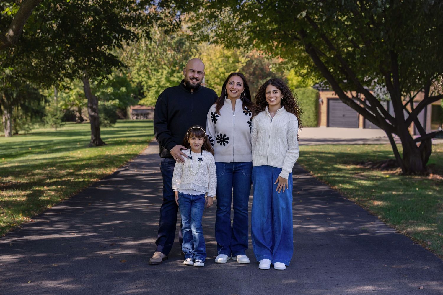 Family of four posing on a tree-lined path in a park during autumn, wearing casual white and dark outfits.