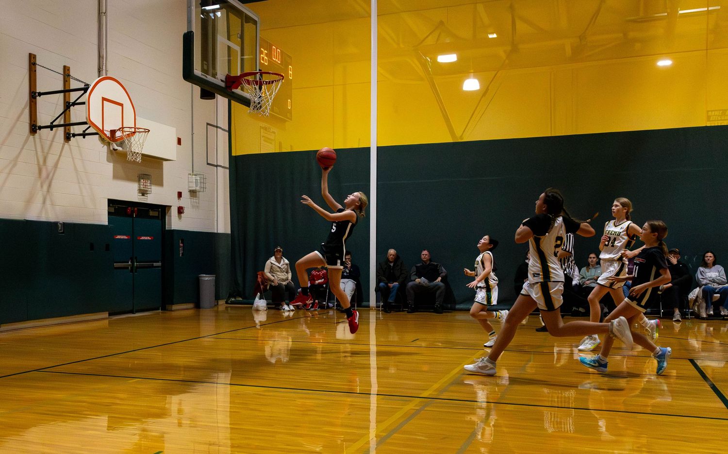 Women's basketball game in indoor gym, player shooting ball toward hoop while teammates and opponents watch.