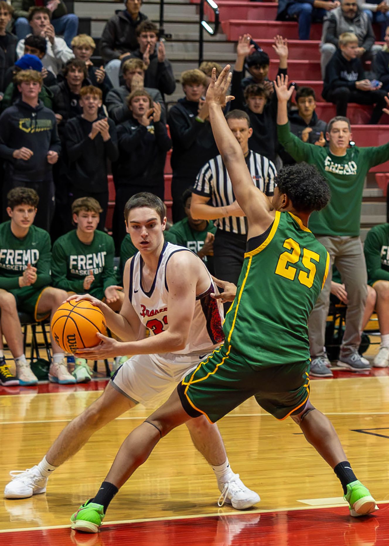High school basketball player in white uniform drives against defender #25 in green during an indoor game.