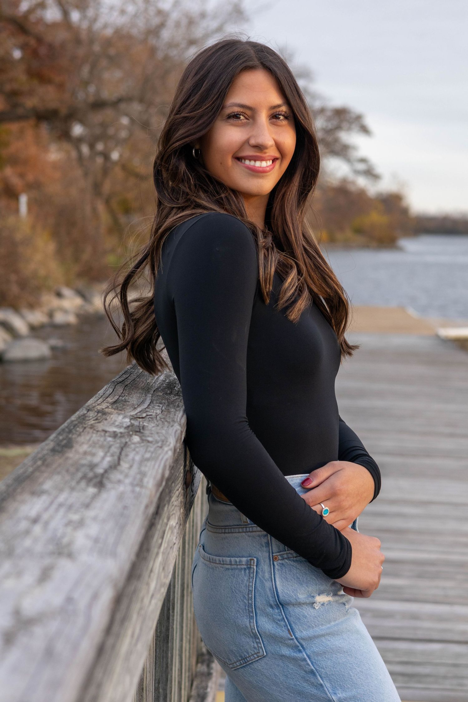 Smiling woman in black long-sleeve top and jeans leaning on wooden bridge railing by a lake in autumn.