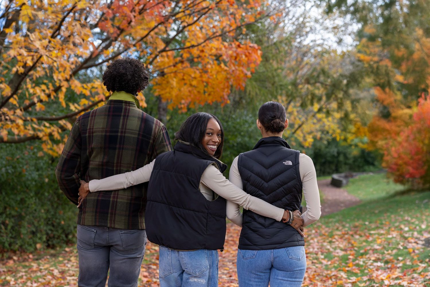 Three friends walk through a colorful autumn park, arms around each other, surrounded by vibrant orange and red fall foliage.