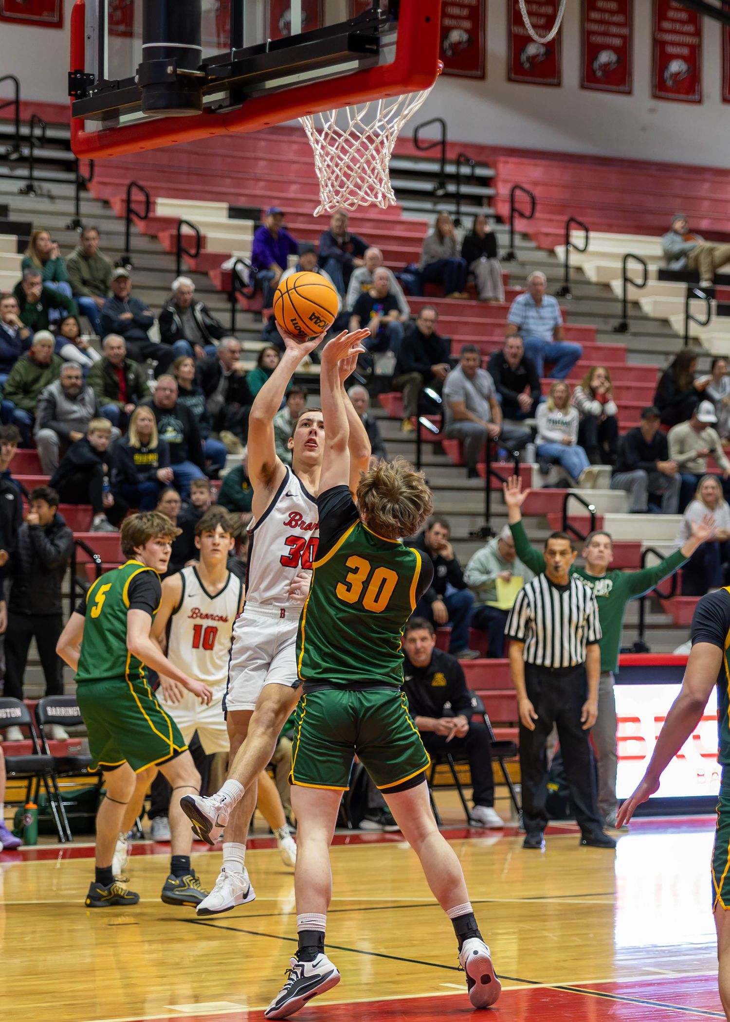 High school basketball player in white uniform shoots over defender in green jersey number 30 during packed gym game.