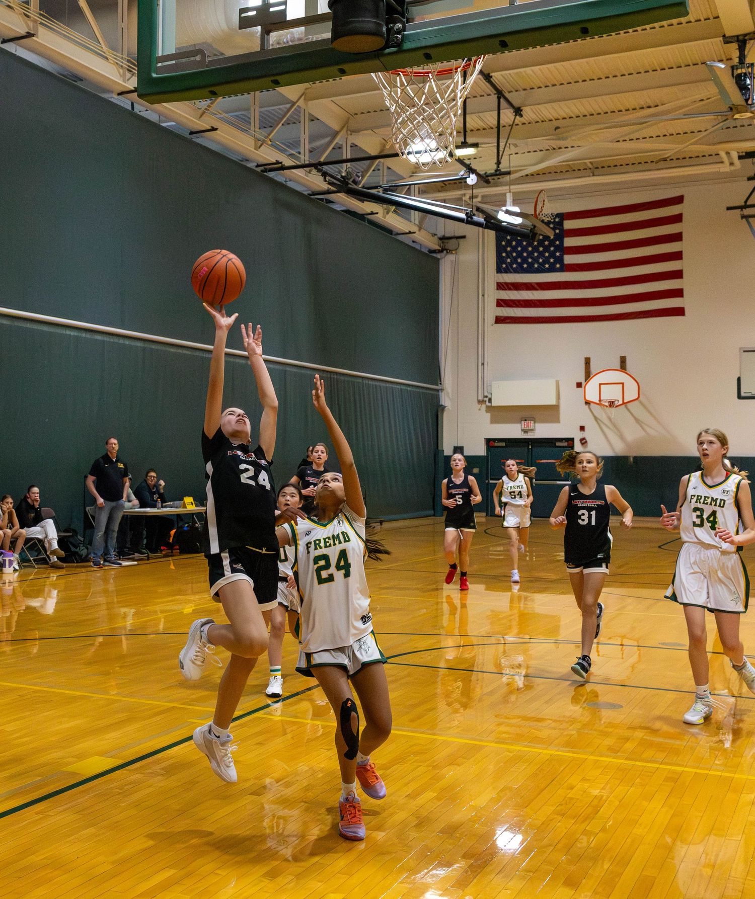 Player #24 in black shoots a layup over a defender during a girls high school basketball game in a green gymnasium.