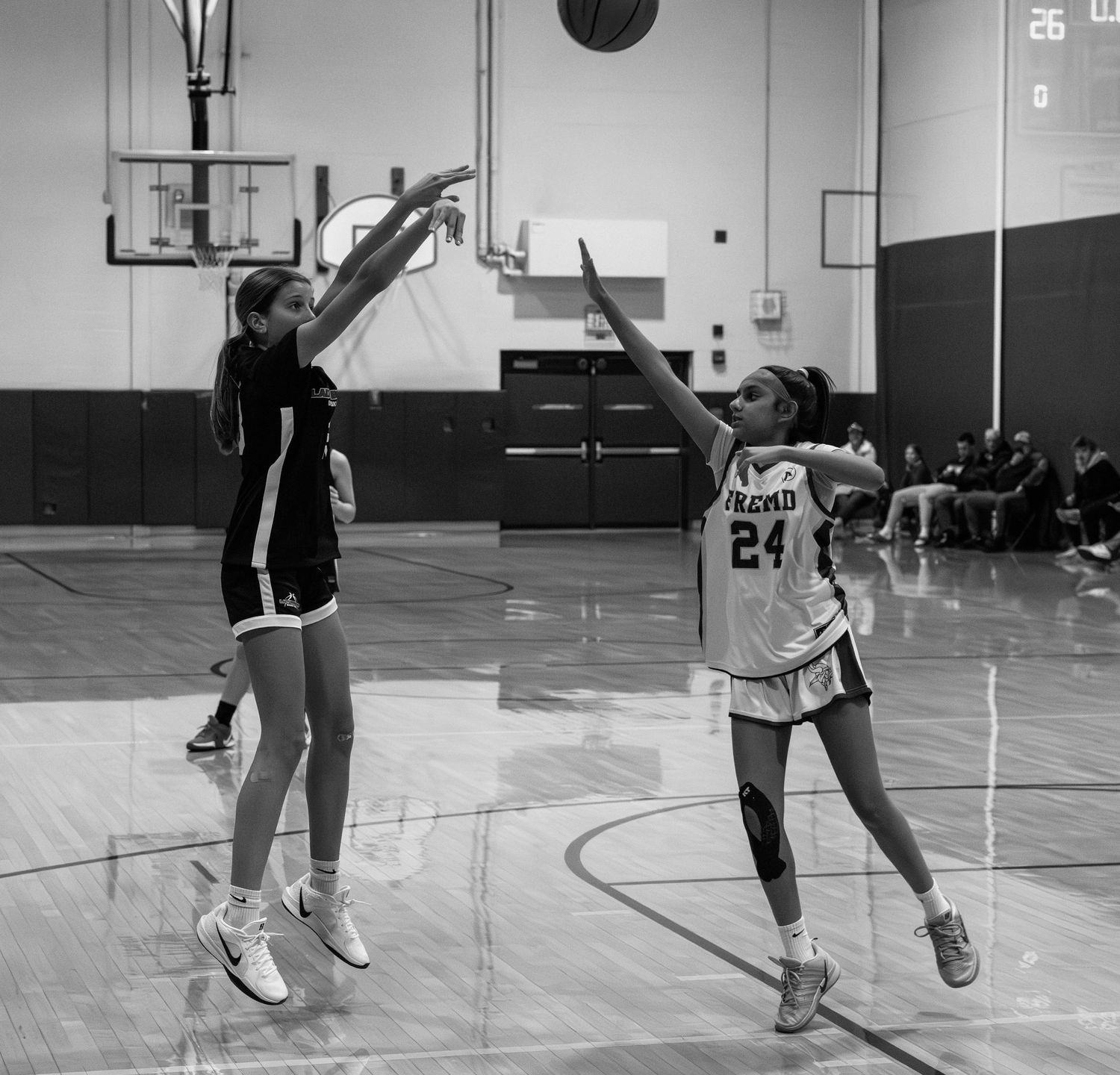 Two female basketball players compete in a black and white photo, one shooting while the other defends during a gym game.