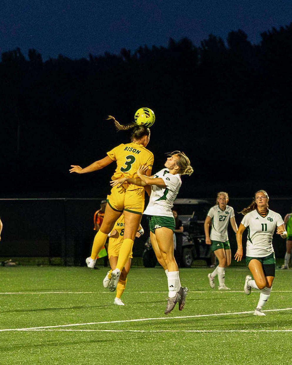 Soccer players compete for a header during an intense nighttime match on a well-lit field.