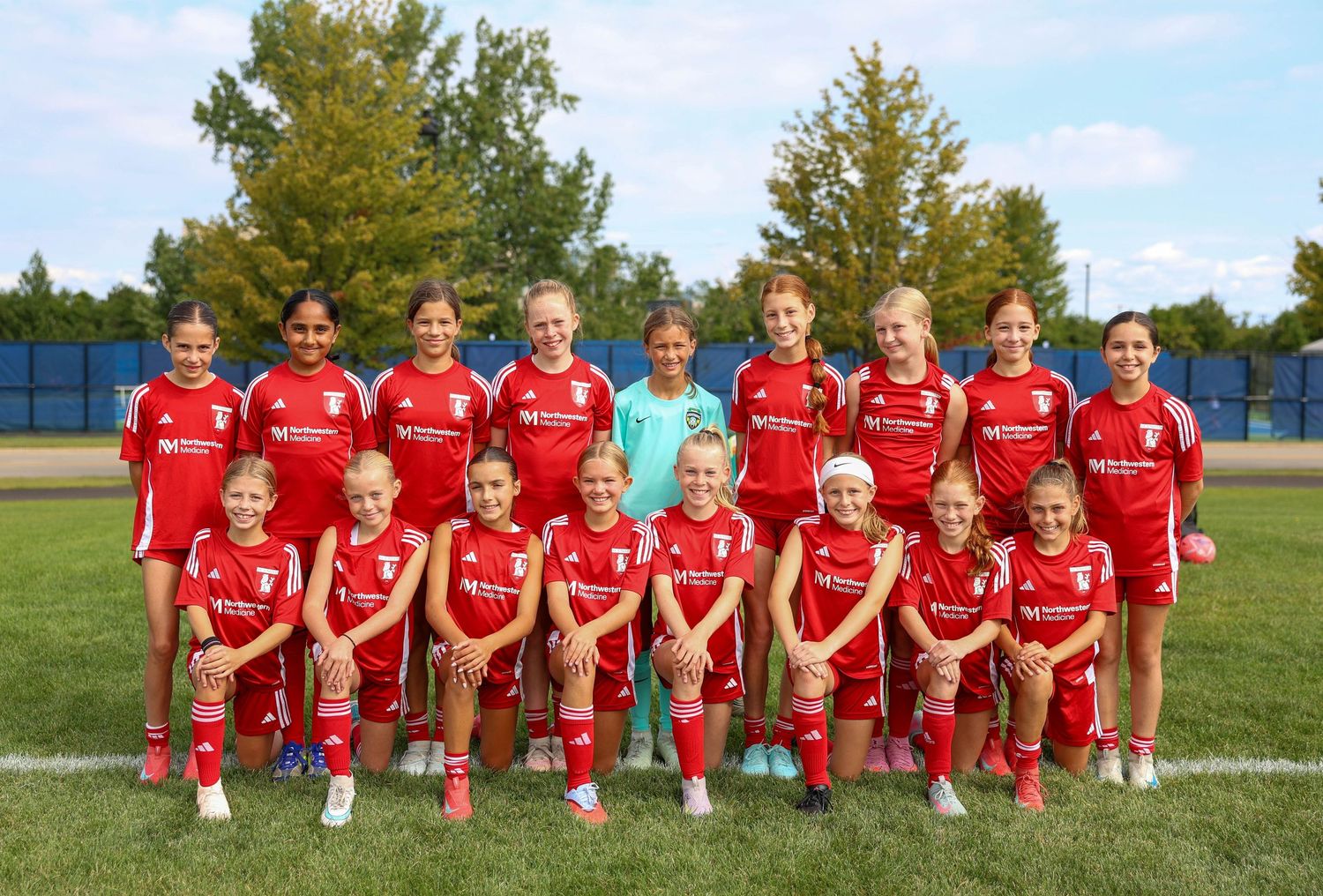 Soccer team members in red jerseys line up on field for team photo with coaching staff.