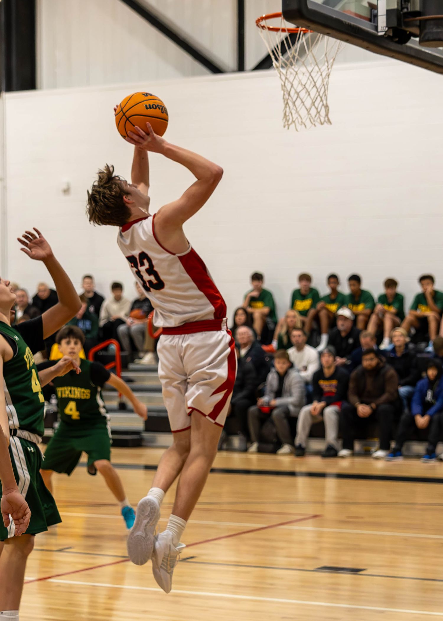 Player #23 in white uniform leaps for a layup during a high school basketball game against green-uniformed Vikings team.