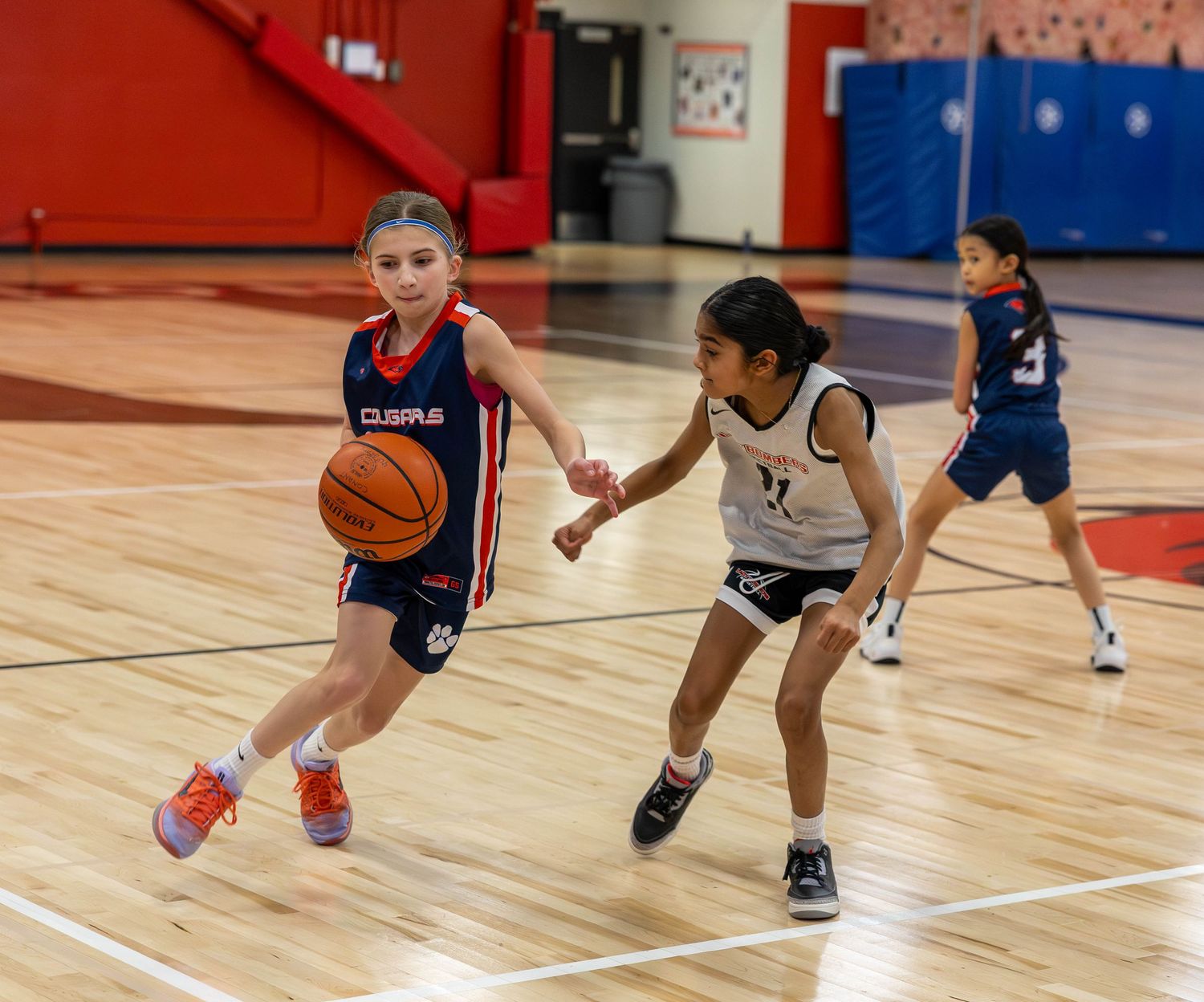 Young girls playing youth basketball in a gym, with one player dribbling past a defender during a game.