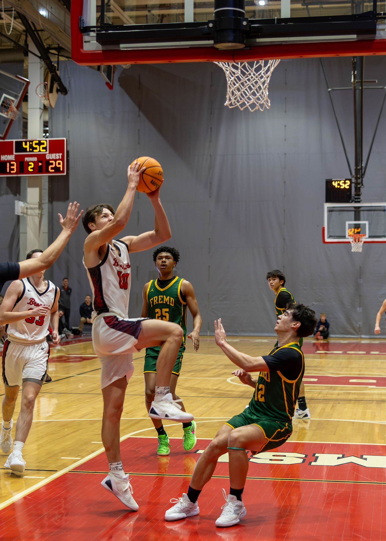 High school basketball player in white uniform drives to the hoop against green-uniformed defenders in a red gymnasium.