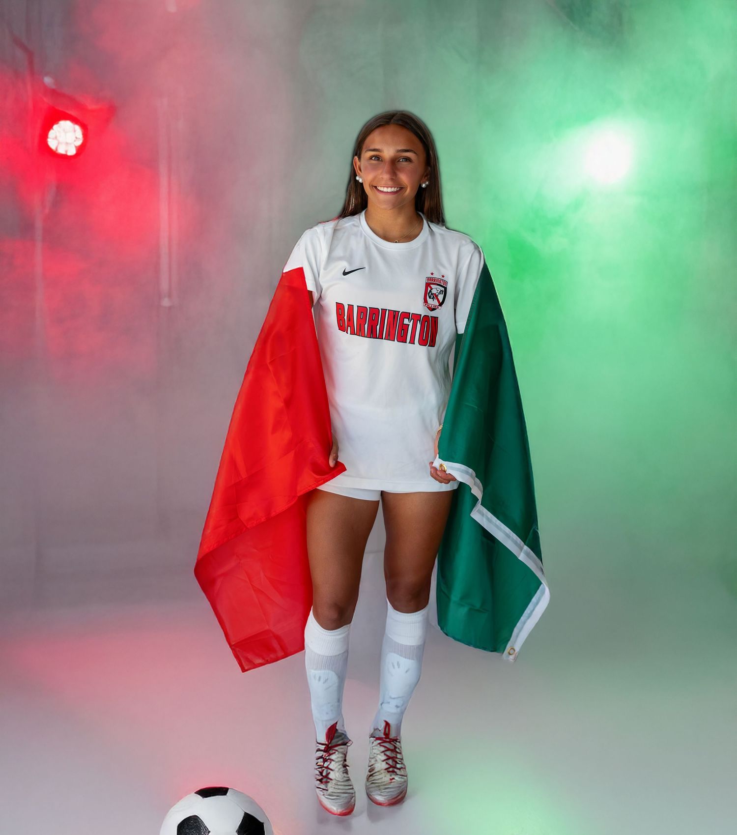 A person in white clothing holds red and green flags while posing against a colorful smoke-filled backdrop.