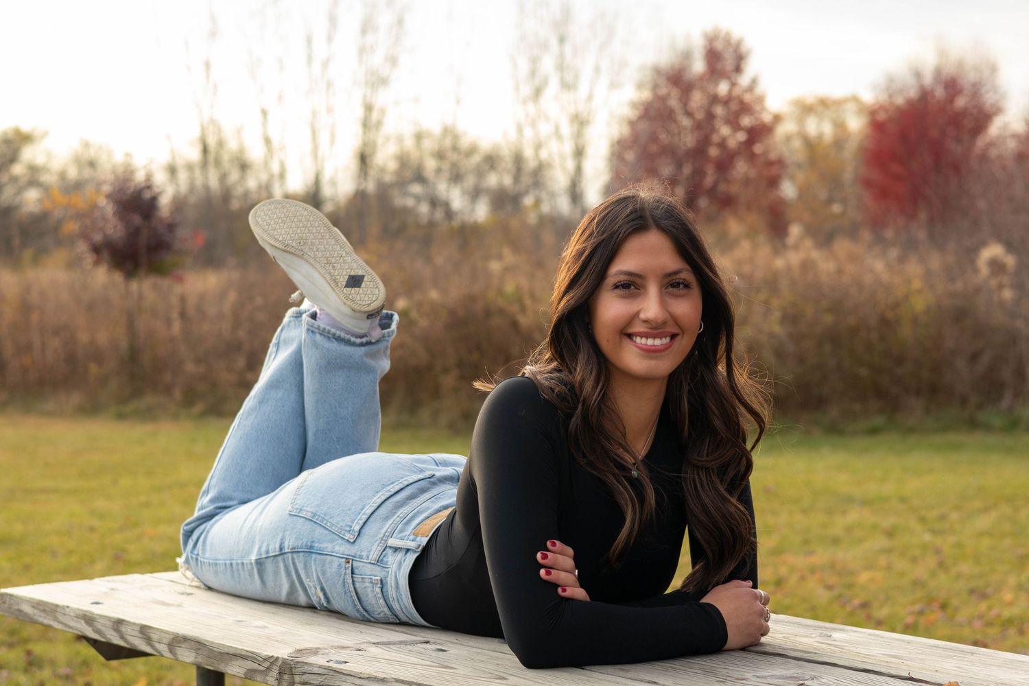 Relaxed outdoor senior portrait on wooden bench with fall foliage in background.
