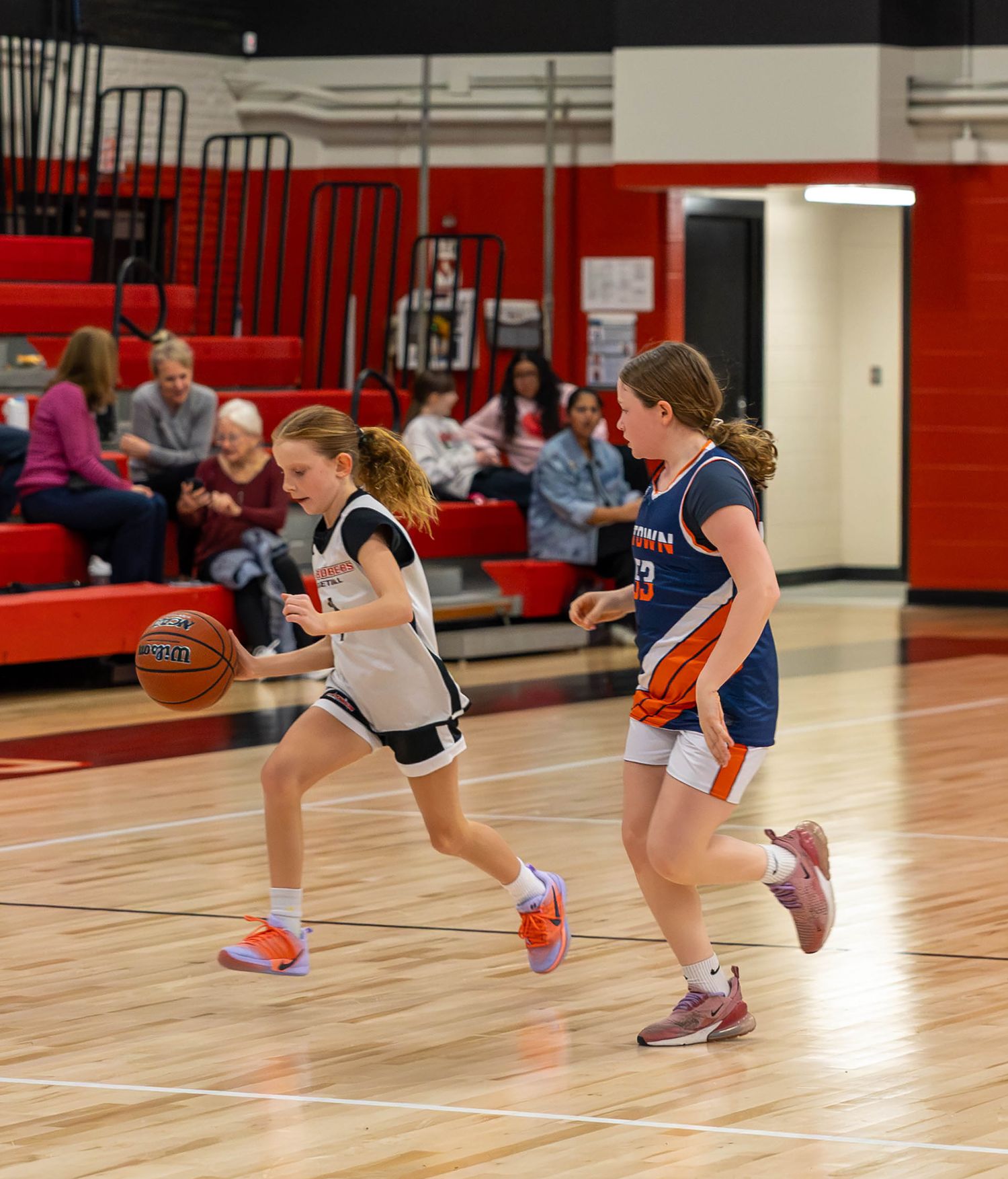 Two young girls playing youth basketball, one dribbling past a defender in a red gymnasium.