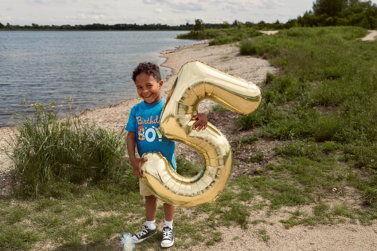 Young child smiling on a beach holding a large gold number 5 balloon, celebrating a 5th birthday outdoors.
