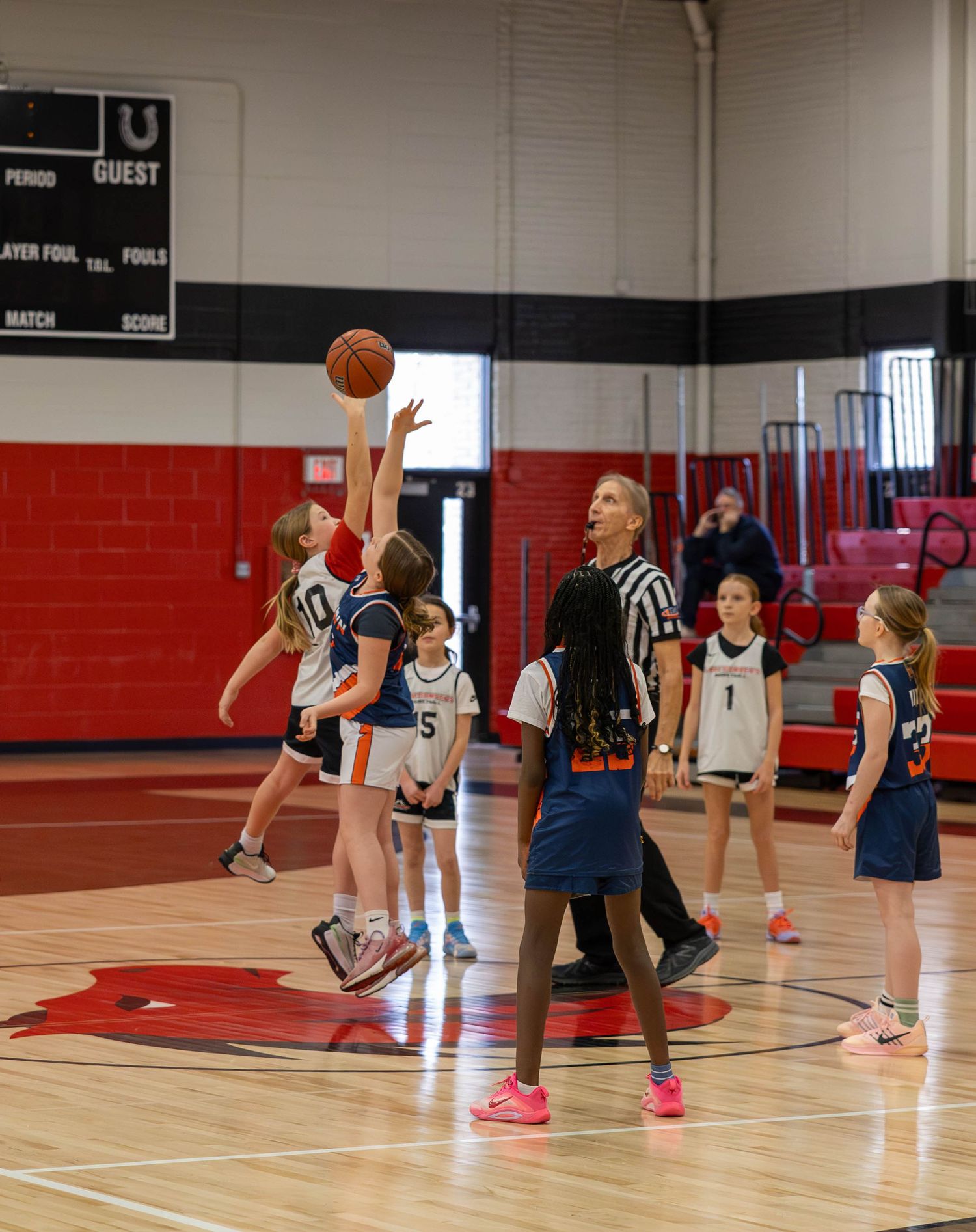 Young girls playing basketball during a tip-off in a red gymnasium with a referee overseeing the game.
