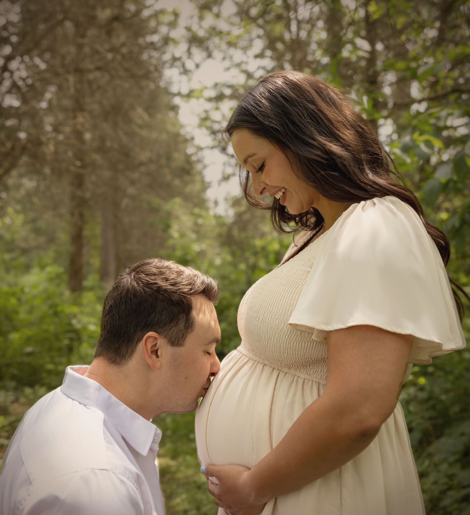 A man kisses a pregnant woman's belly during an outdoor maternity photo shoot surrounded by lush green trees.