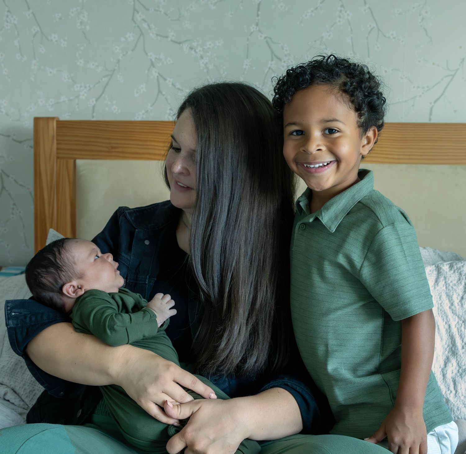 A woman with long dark hair sits on a bed holding a newborn while a smiling young boy in green sits beside her.