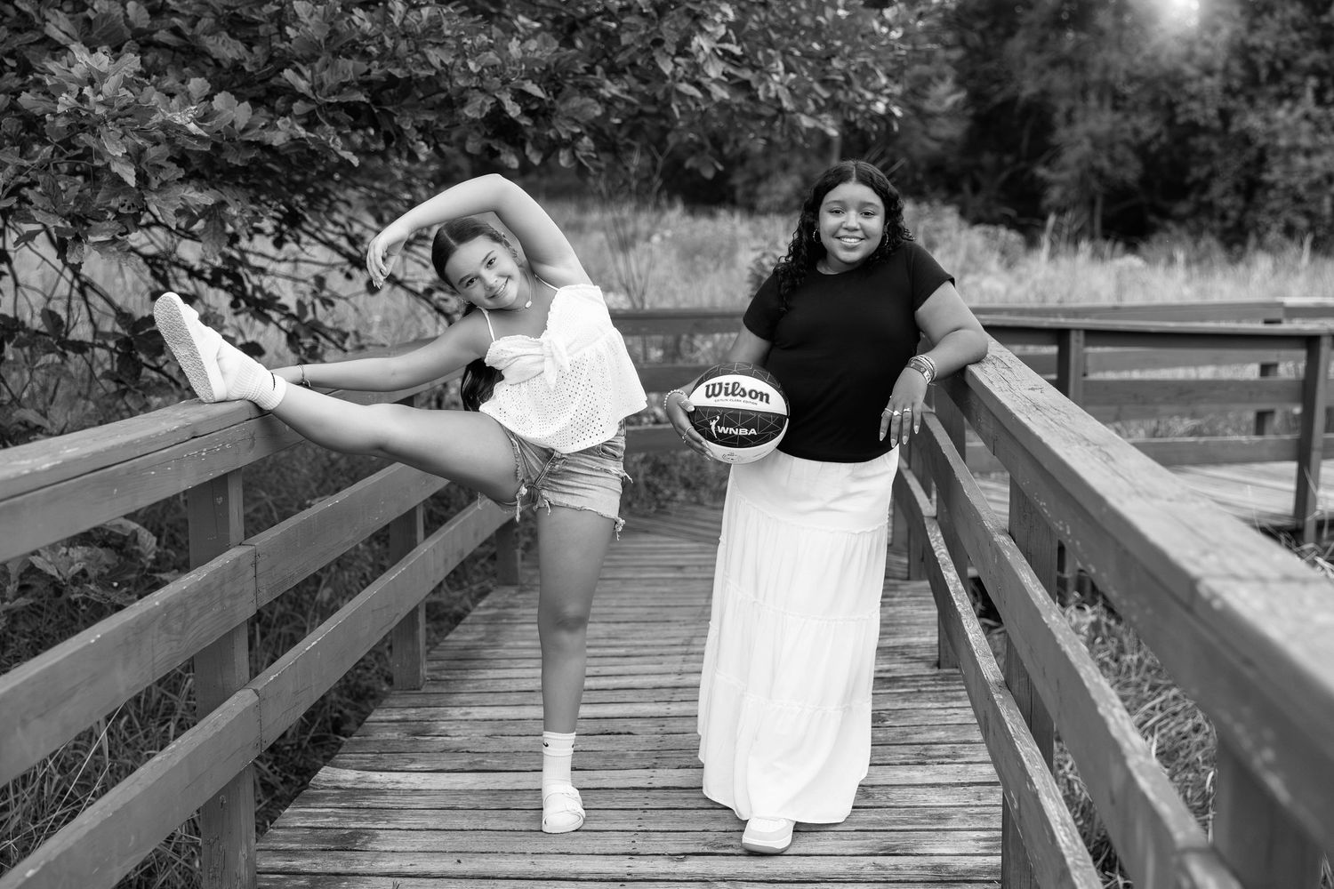 Two women laugh and pose playfully on a wooden bridge while holding a picnic basket during a summer afternoon.