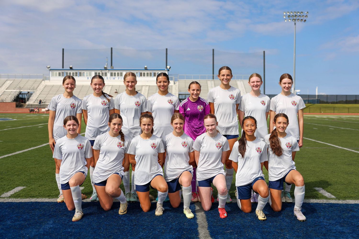 Female athletes from a soccer team pose together in front of a goal on a sunny day with blue skies.