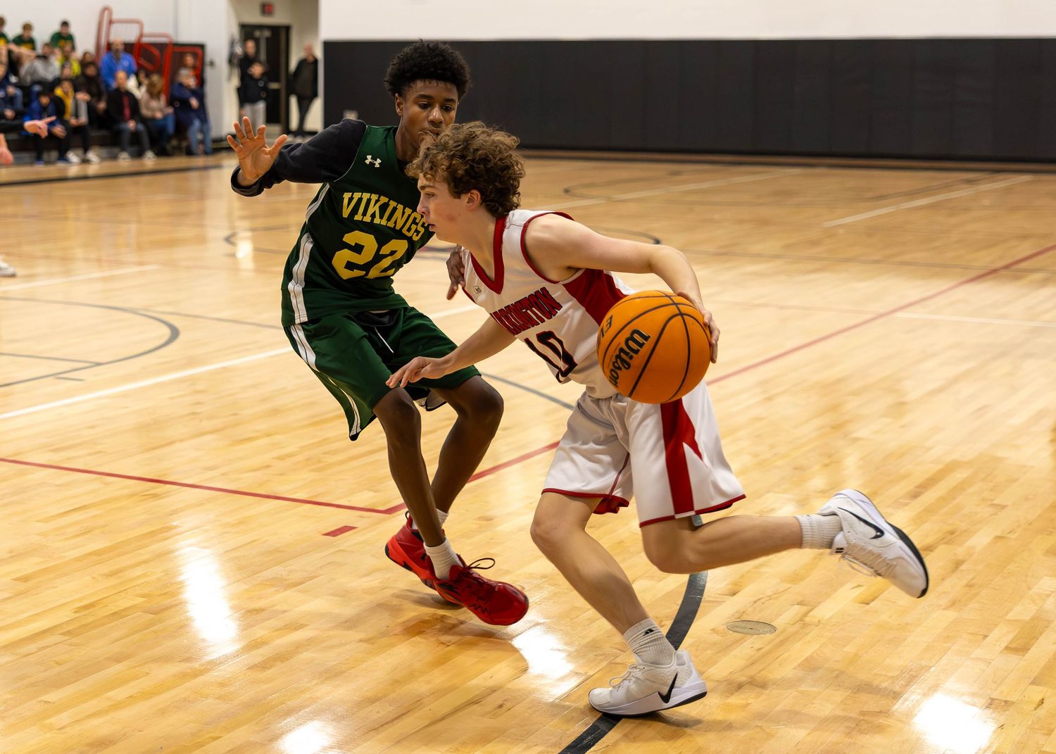 High school basketball player in red dribbles past defender in green Vikings jersey during indoor game.